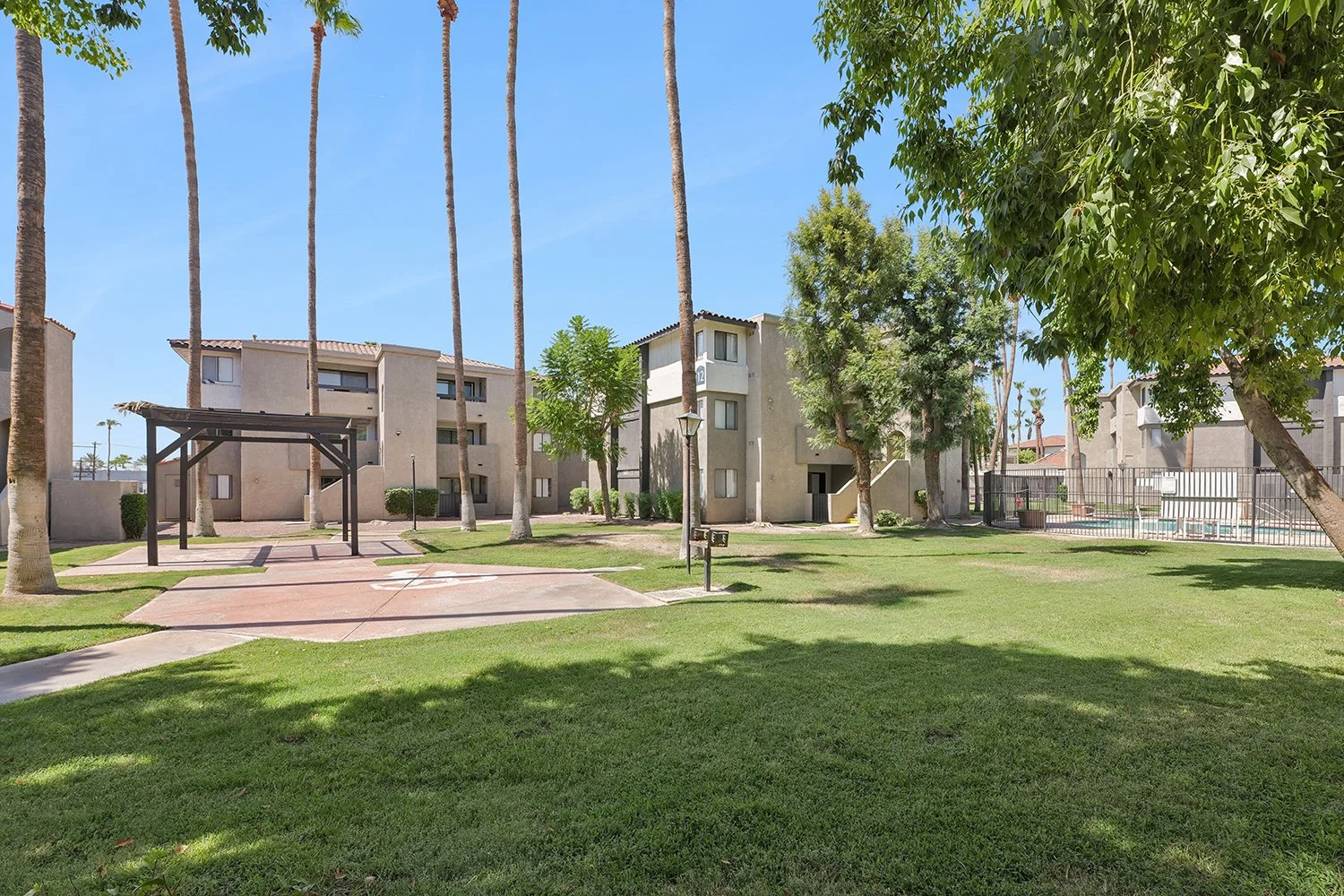Included in this image is a spacious grass lawn within the apartment's courtyard. On the left sits a dark wooden pergola covering a concrete patio, while a fenced-in swimming pool area is in the right background. 
