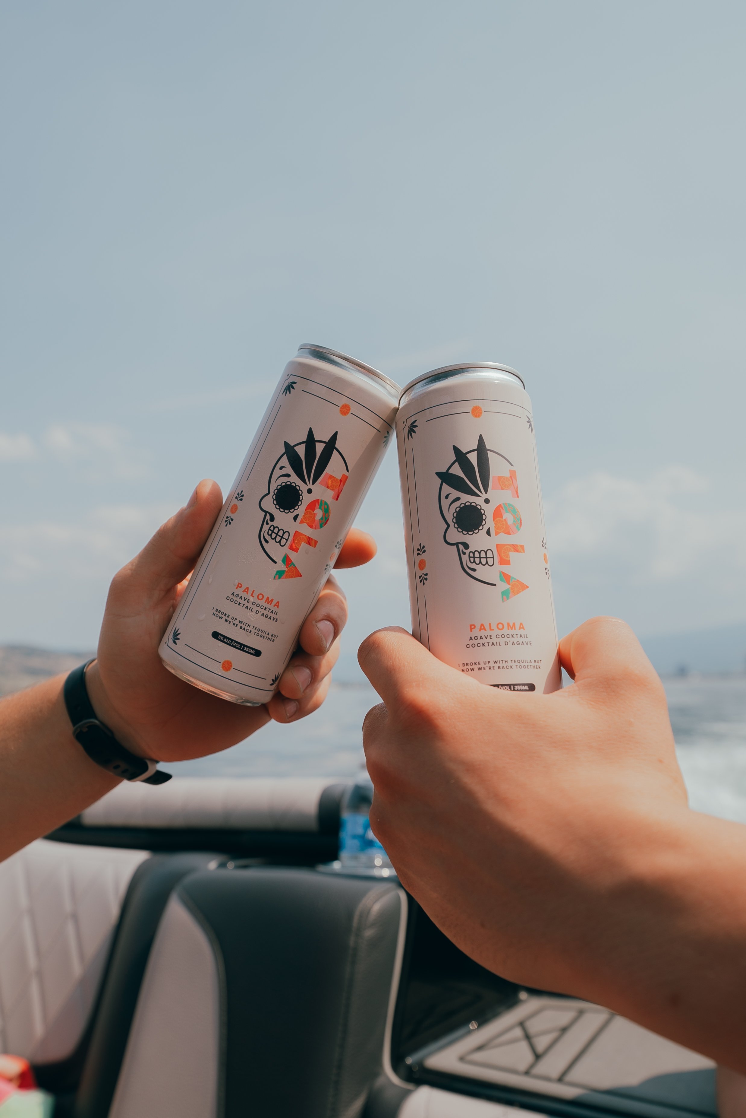 Two cans of Paloma agave cocktail being held up towards the sky in a toast, with a background of a clear blue sky and a part of a vehicle interior visible.