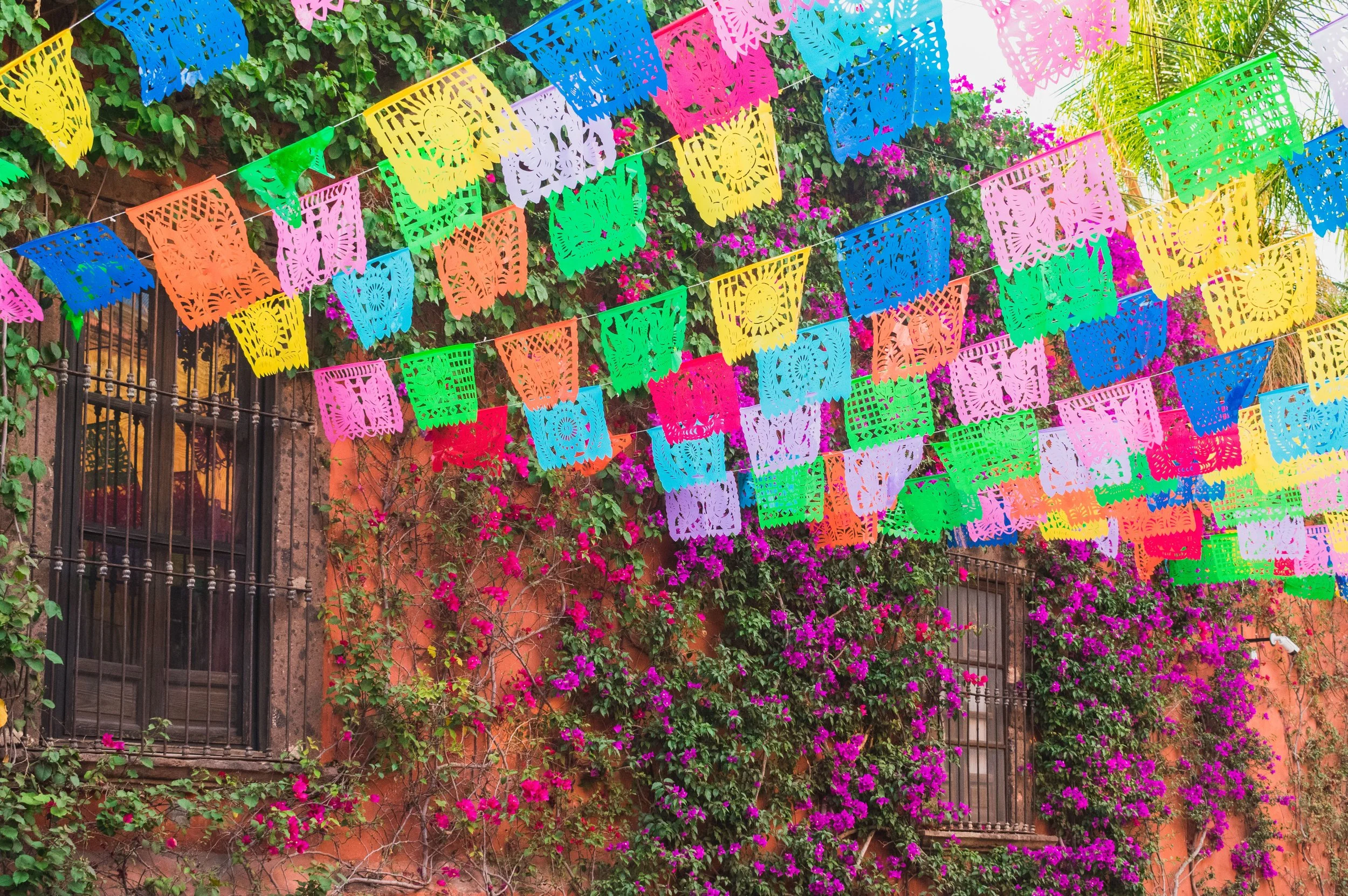 Colorful papel picado banners hanging above a flower-filled outdoor area with a brick wall and iron fence.