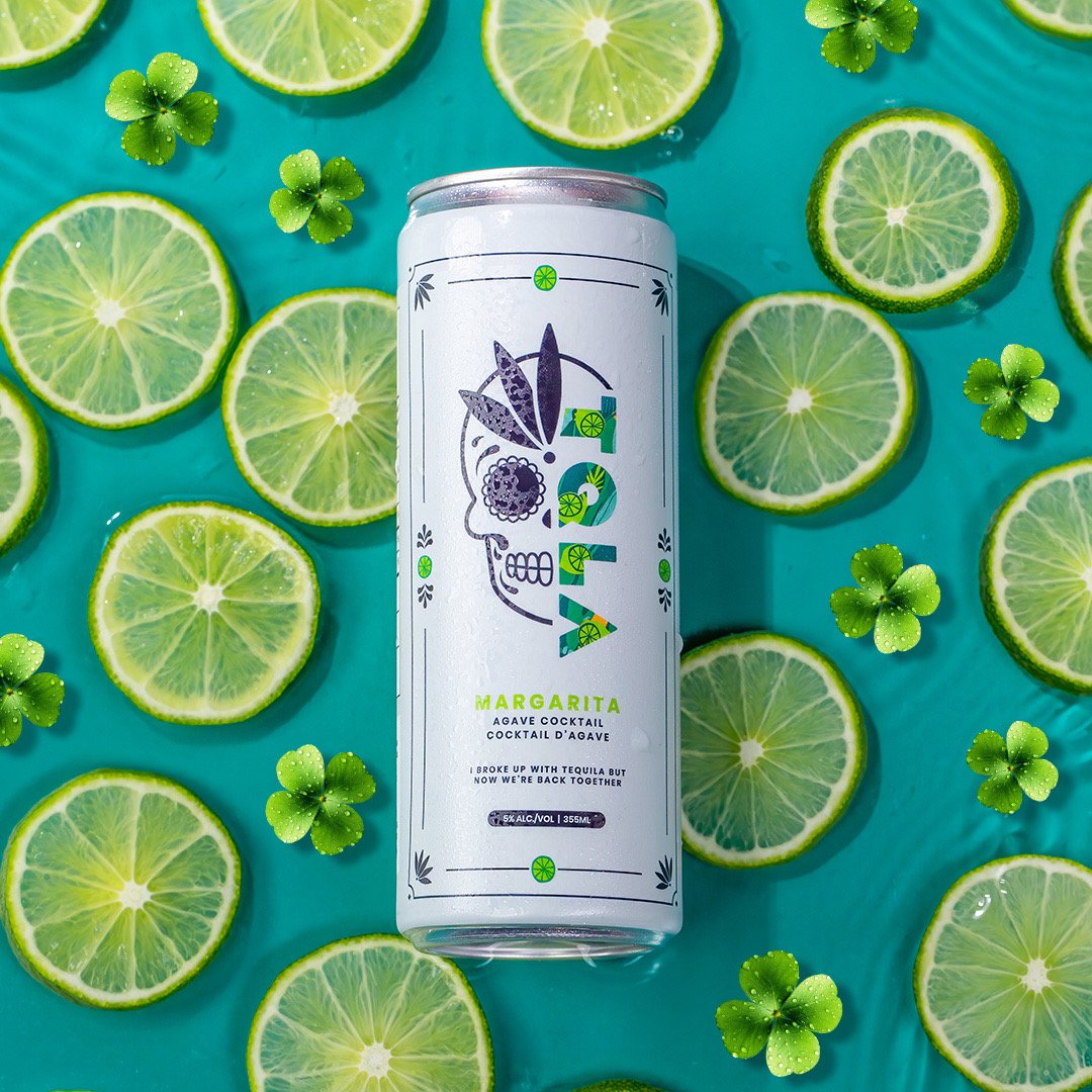 A can of AVA Mango Margarita cocktail surrounded by lime slices and small shamrock-shaped leaves on a turquoise background.