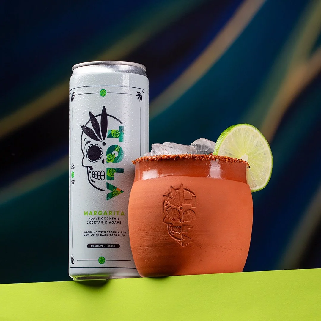 A can of agave margarita cocktail and a ceramic cup with a lime wedge on the rim, filled with ice, against a dark background.