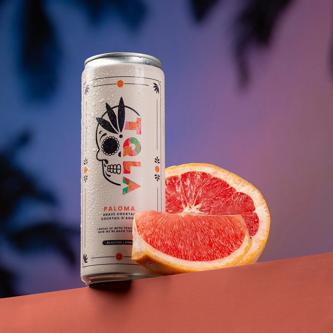 A can of Paloma agave cocktail next to a halved grapefruit on a pink surface with a colorful blurred background.