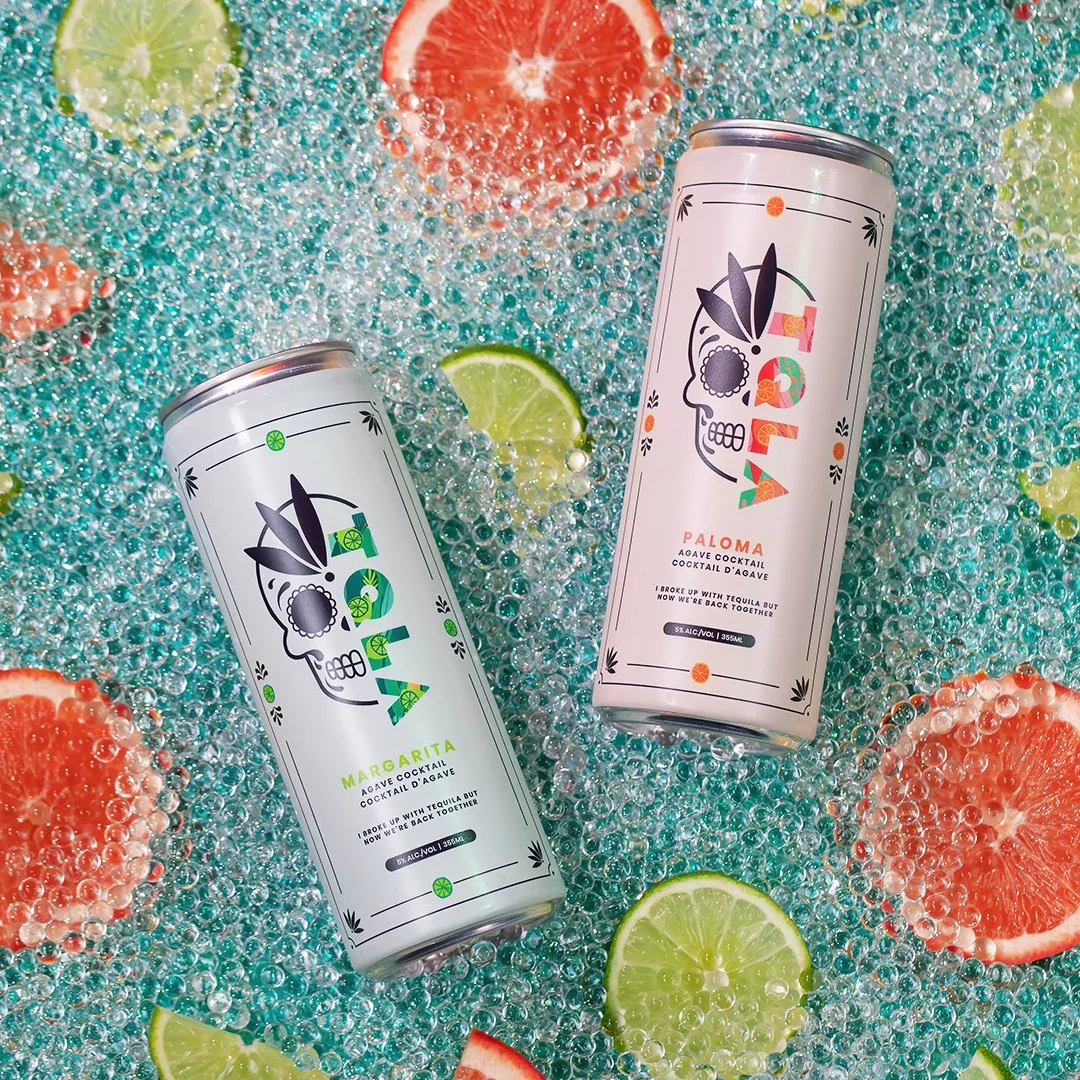 Two cans of cocktail drinks, one labeled Margarita and the other Paloma, are placed on a surface surrounded by sliced limes, grapefruits, and bubbles. The cans have a modern skull design with floral and abstract elements.