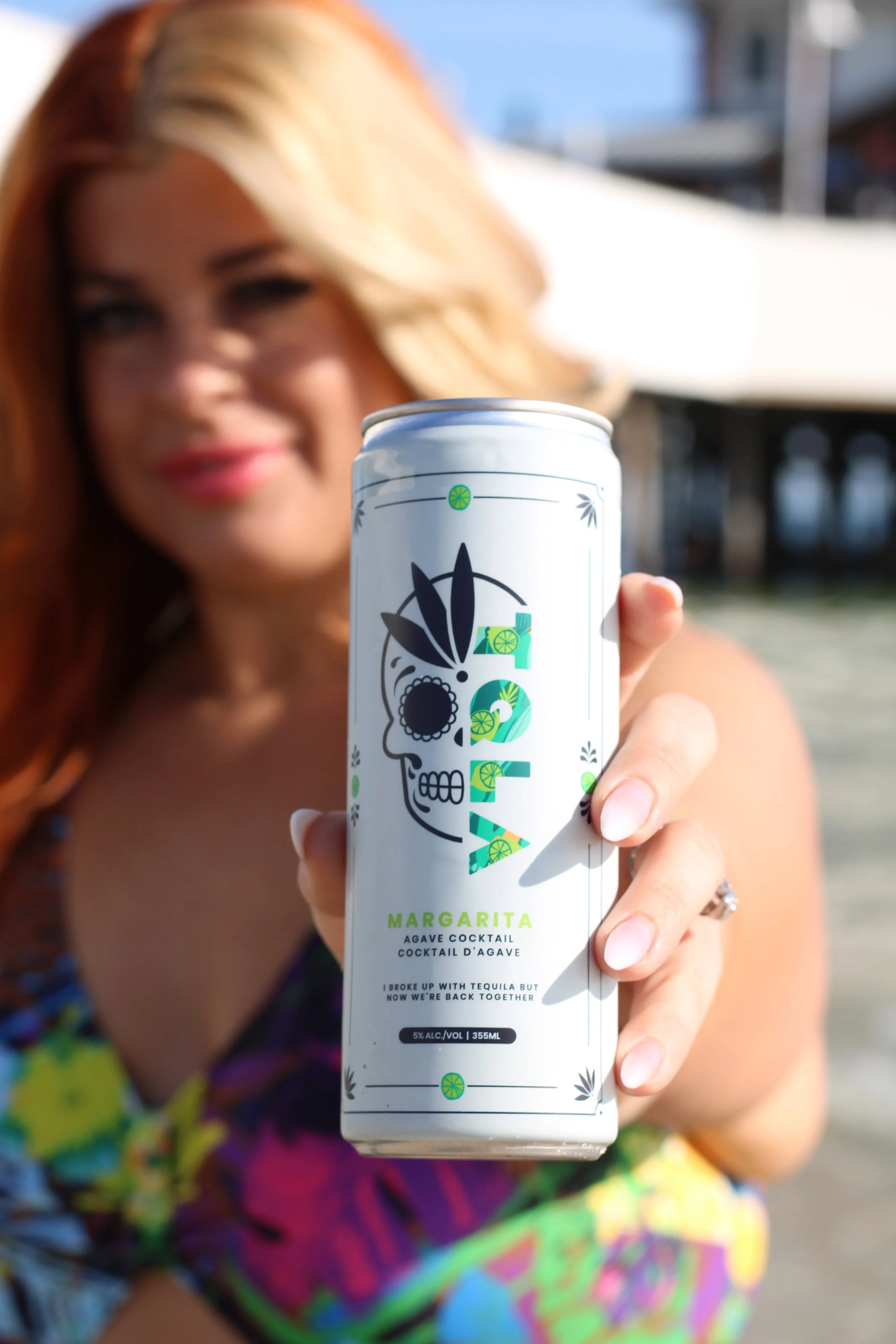 A woman with red hair holding a can of lime-flavored margarita cocktail outdoors, smiling and wearing a colorful dress.