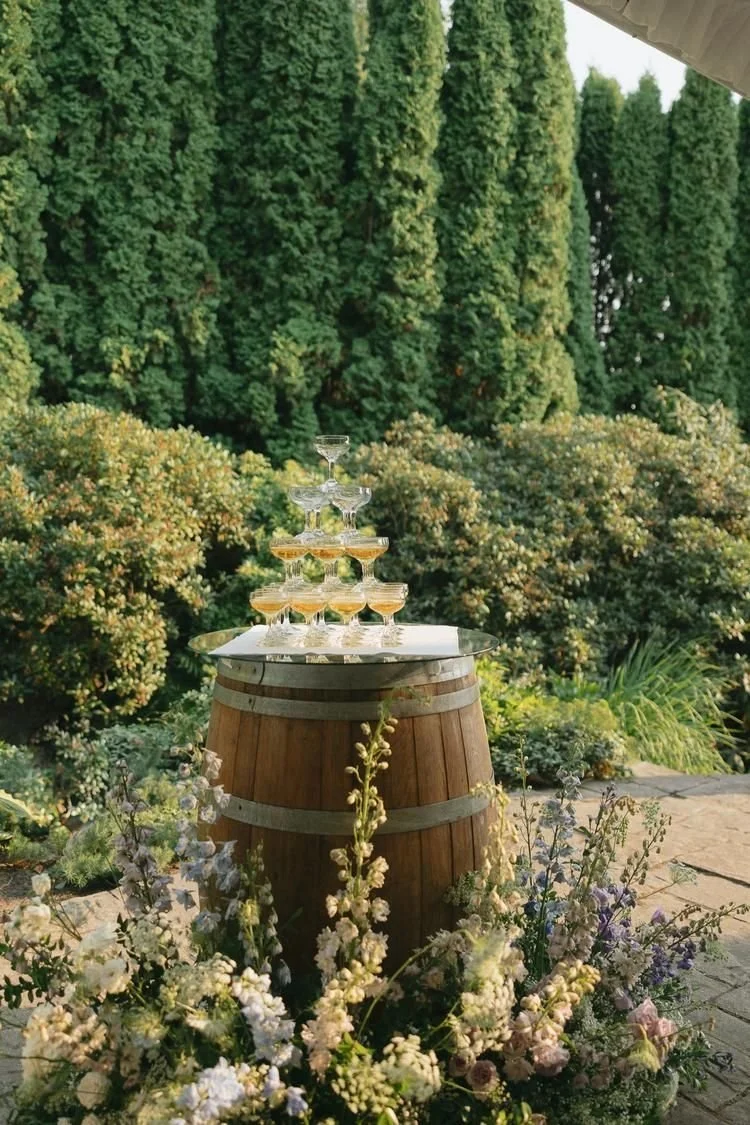 A champagne tower on top of a wine barrel with an arrangement of delphinium, hydrangea and lisianthus. Niagara wedding flowers