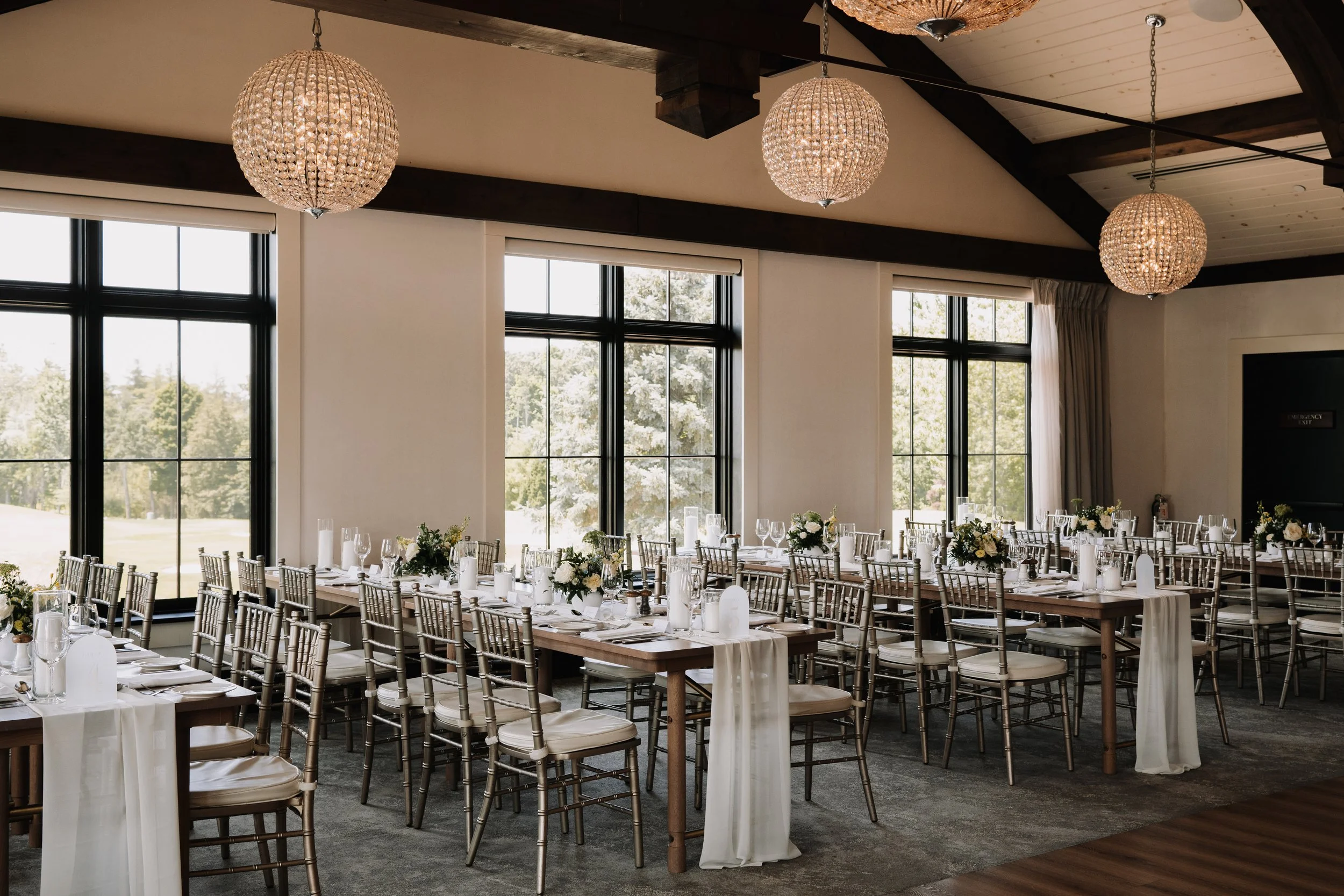 Elegant indoor wedding reception with long wooden tables, white flowers, candles, and chairs with rattan backs.