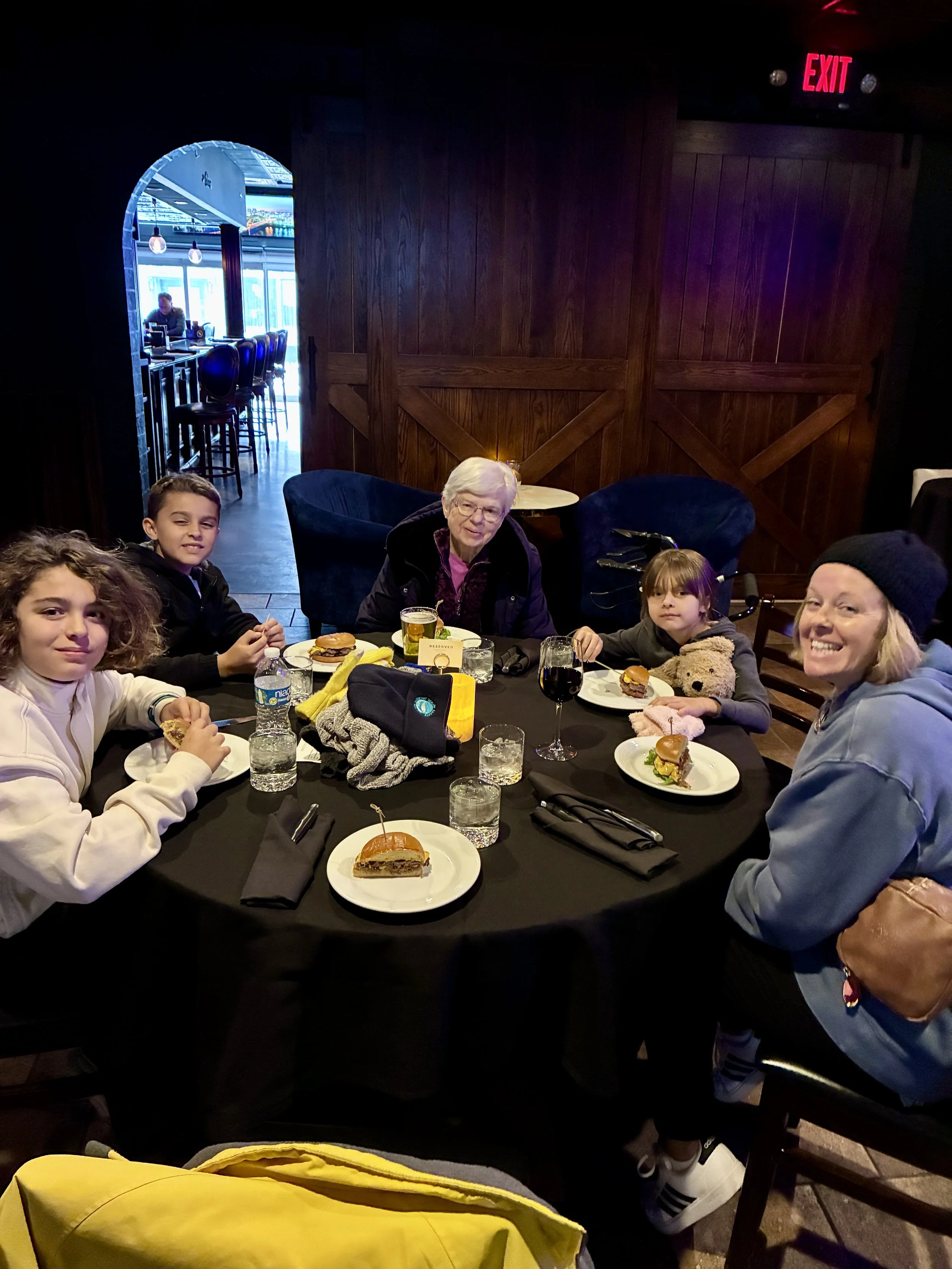 A family of five sitting at a restaurant table with burgers and drinks, smiling for the photo. There are three children, two women, and various drinks on the table, with a dark wooden and cozy interior background.