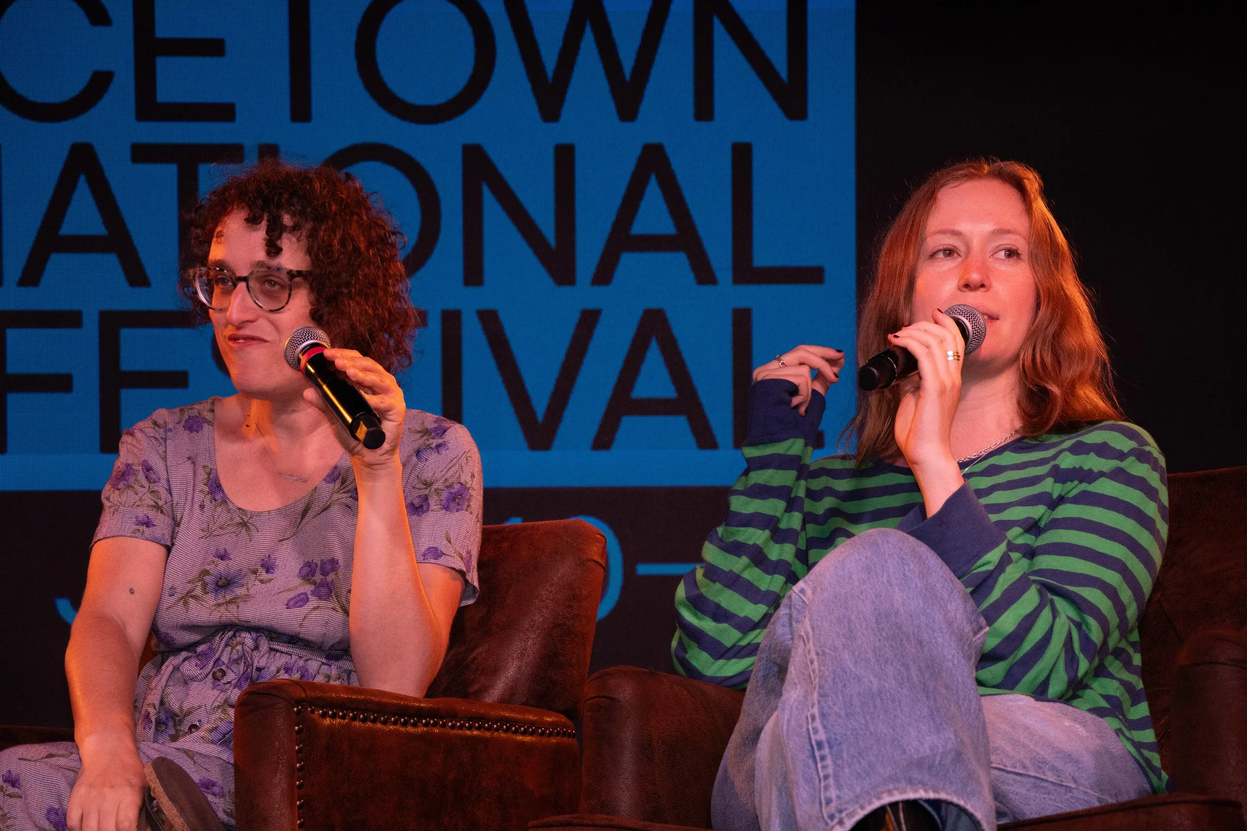 Two women sitting on stage, each holding a microphone. The woman on the left has curly hair, glasses, and is wearing a floral top, while the woman on the right has long red hair and is wearing a green and black striped shirt. Behind them, there is a 