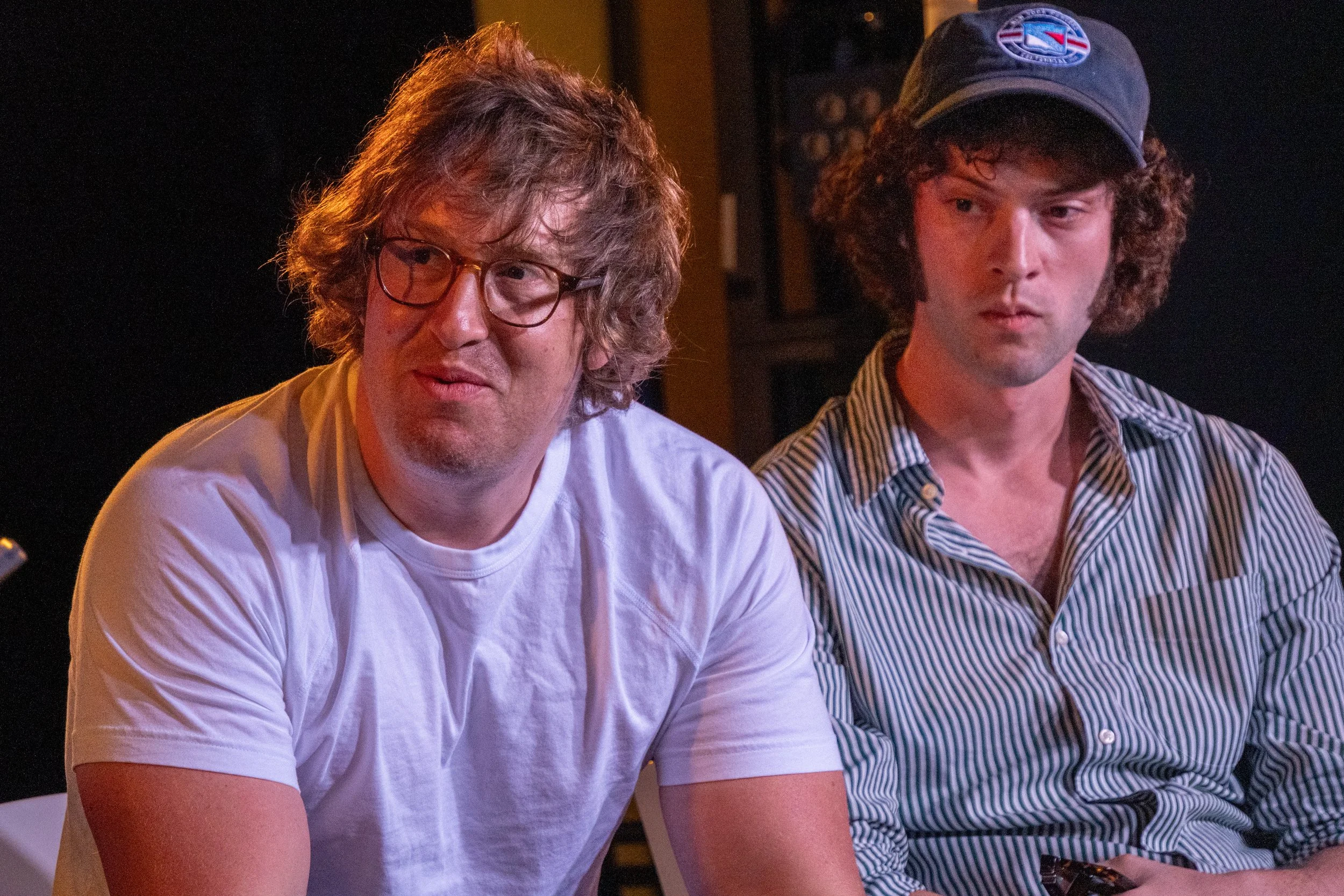Two young men sitting together indoors, one wearing glasses and a white t-shirt, the other wearing a baseball cap and a striped shirt, both looking off to the side with neutral expressions.