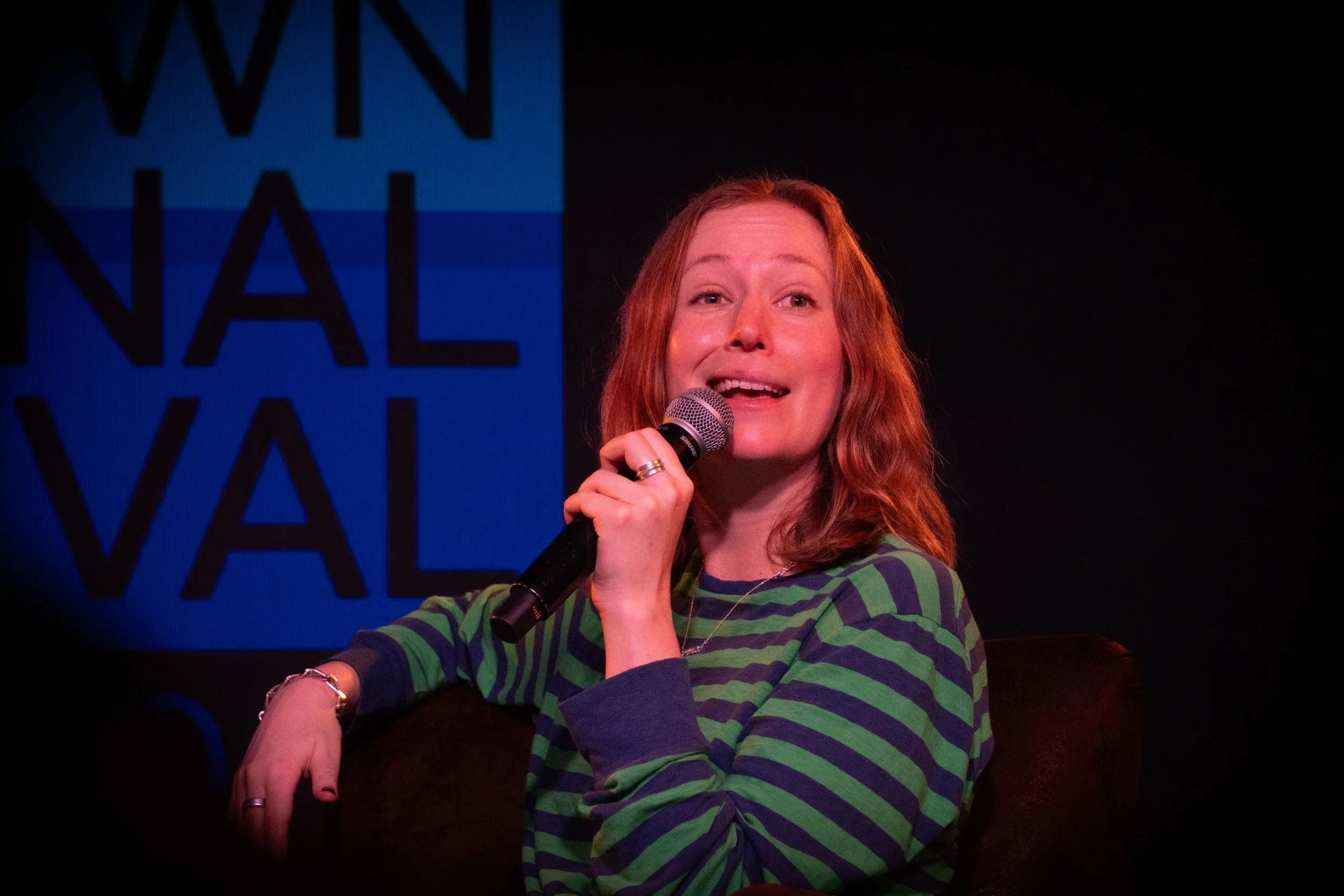 A woman with red hair, wearing a green and purple striped shirt, holding a microphone and smiling on stage with a blue and black background.