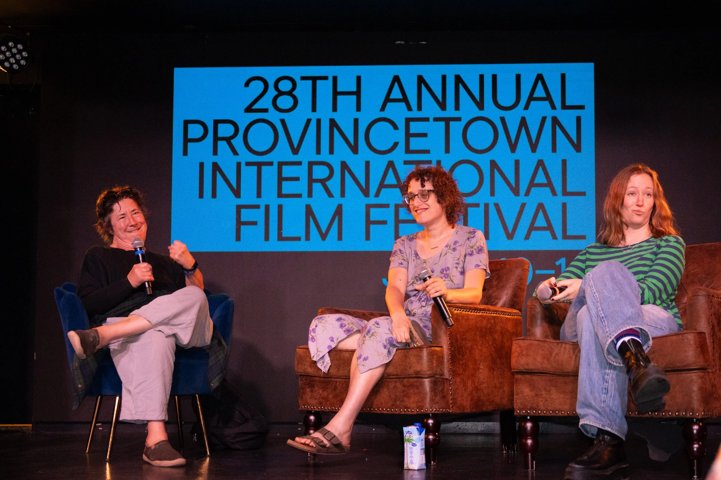 Three women sitting on stage with a large blue sign behind them that reads '28th Annual Provincetown International Film Festival.' The women are engaged in a panel discussion, each holding a microphone, with casual attire and relaxed postures.
