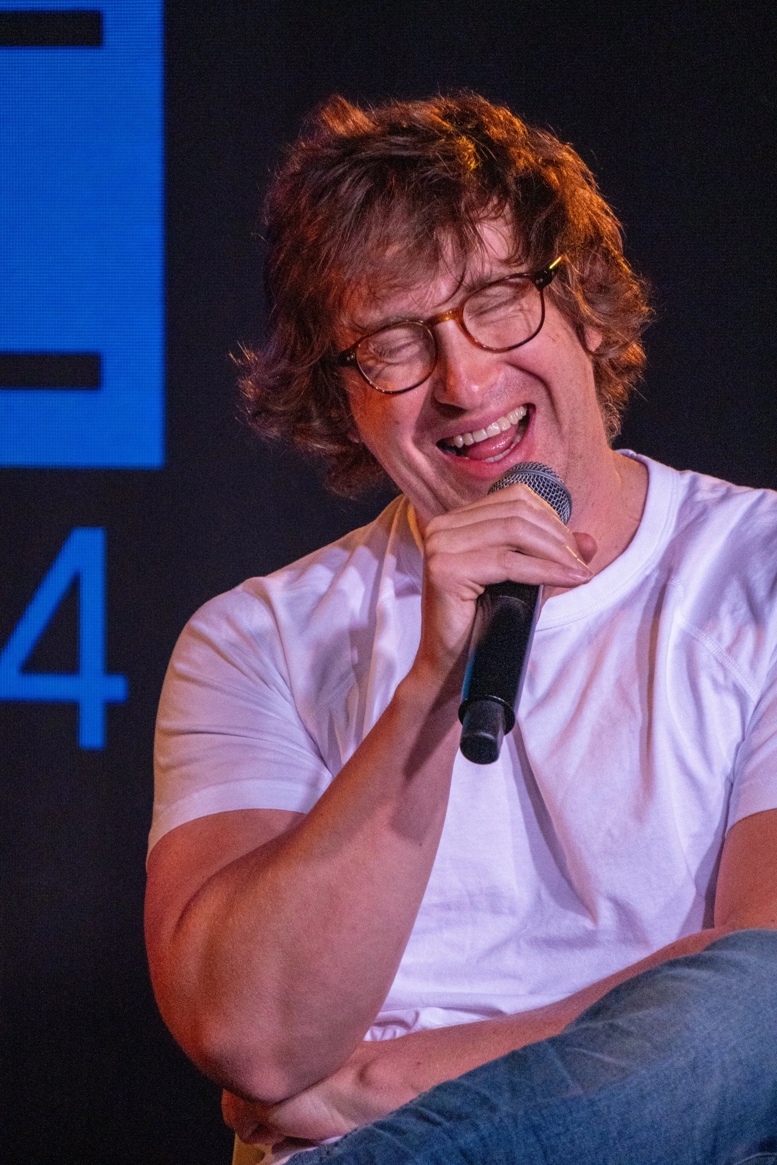 A young man with curly brown hair and glasses is smiling with his eyes closed while talking into a microphone. He is wearing a white T-shirt and sitting with his arms crossed.