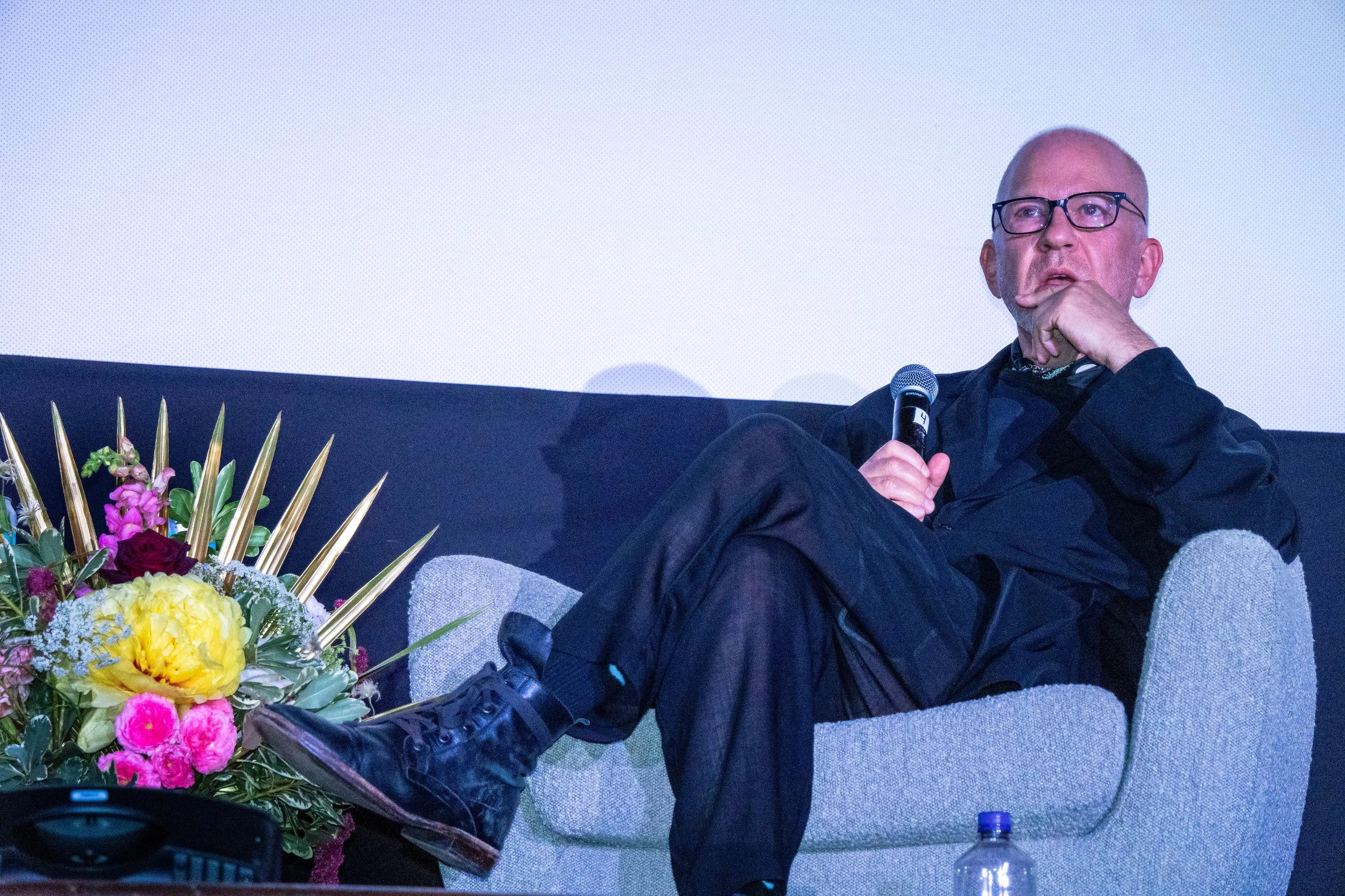 A man with glasses sitting on a chair with his leg crossed, holding a microphone, wearing black clothes, next to a floral arrangement, in front of a plain white background.