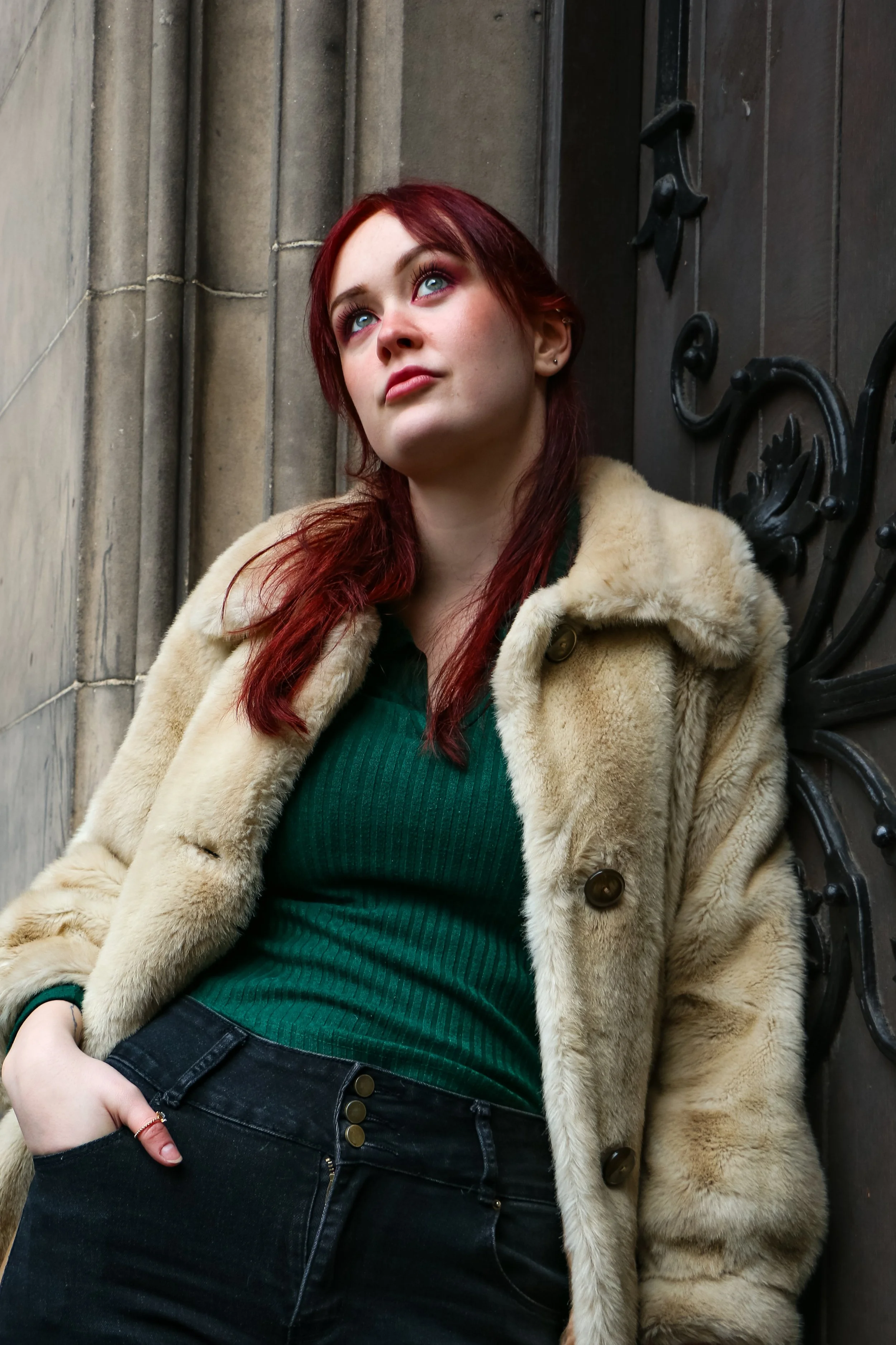 A young woman with red hair and blue eyes sitting against a stone wall and wooden door, wearing a beige fur coat and green top.