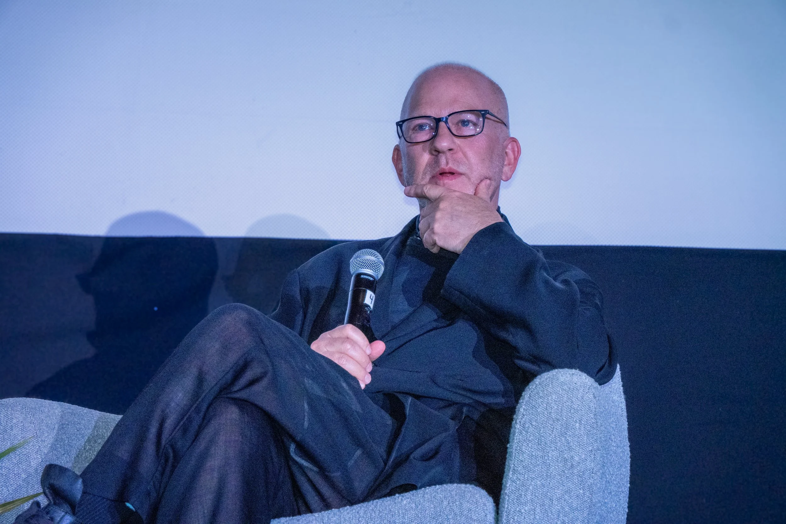 A man with glasses, a beard, and a shaved head sitting on a gray couch with a blanket. He is holding a microphone and resting his chin on his hand, looking contemplative.