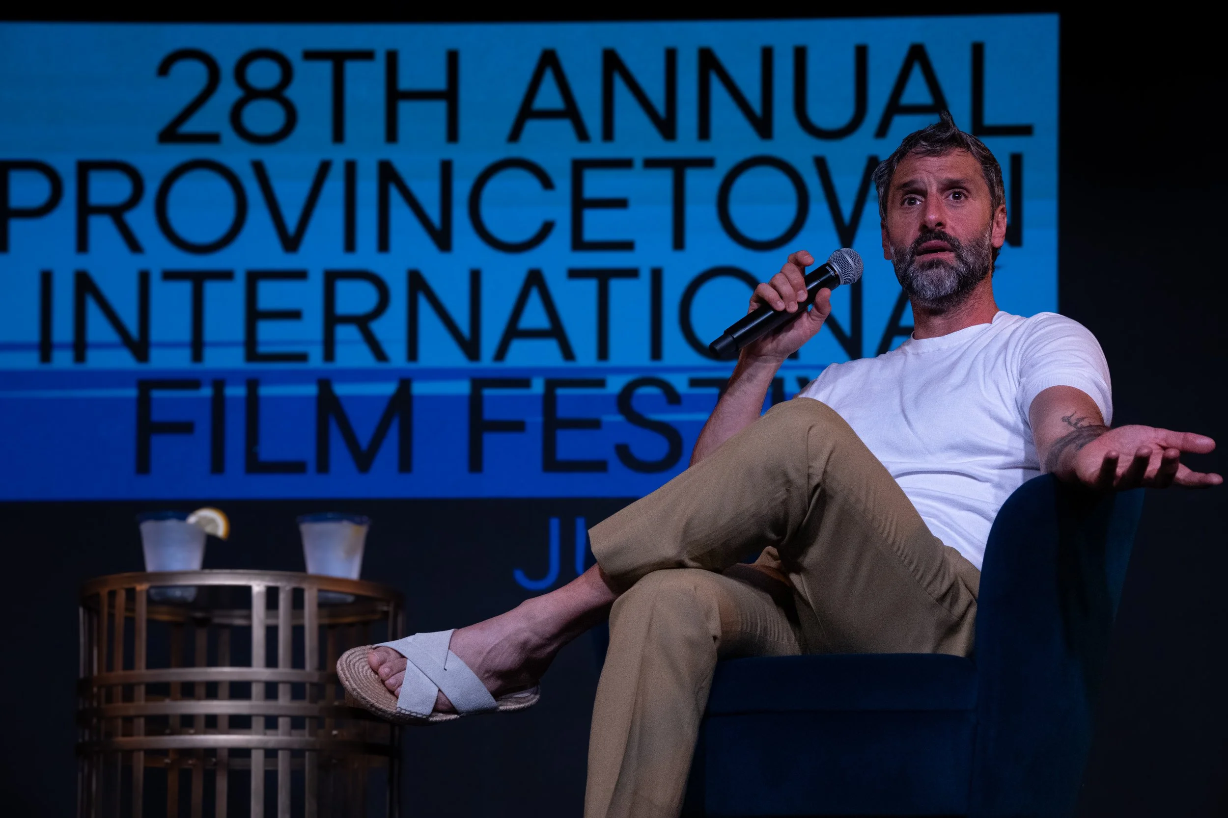 A man with a beard and gray hair sitting on a stage, holding a microphone, with a large blue sign behind him that reads '28th Annual Provincetown International Film Fest'.