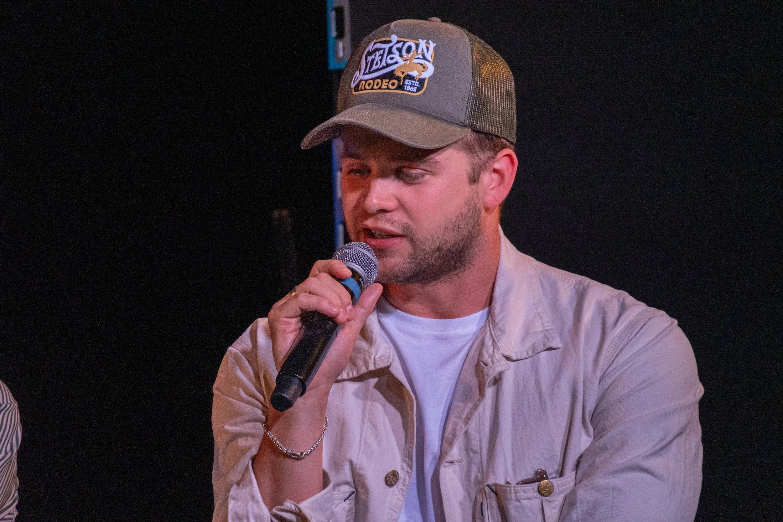 A man wearing a beige jacket and a brown baseball cap with a logo reading 'Weguen Rodeo 1865' is speaking into a microphone against a black background.