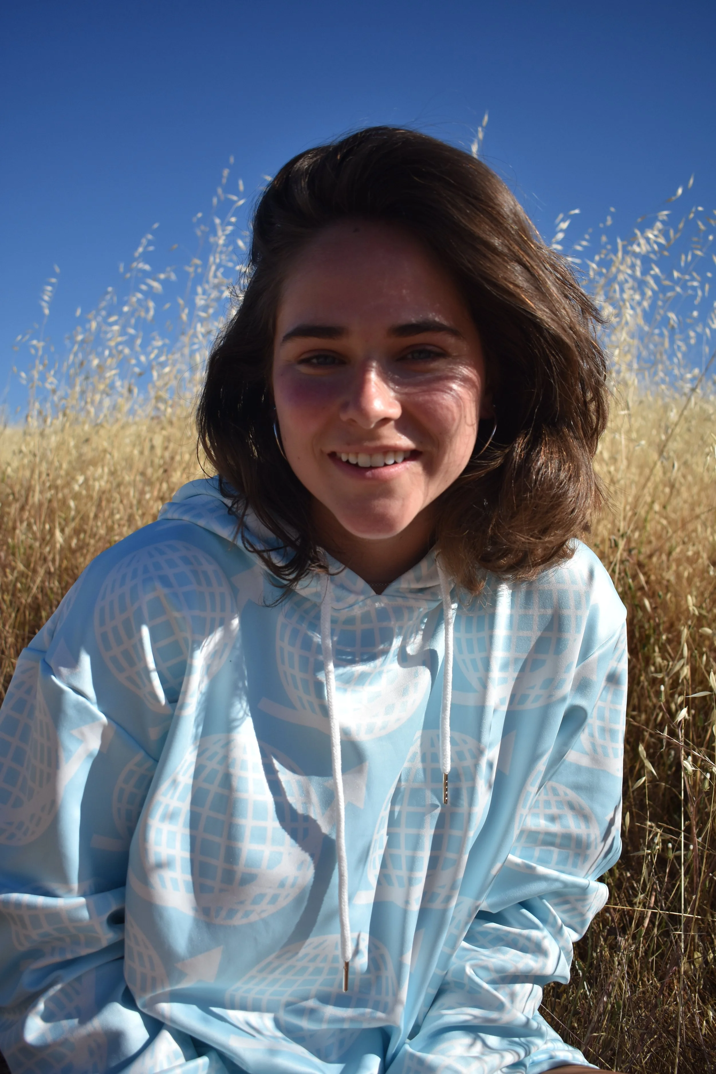 Young woman with dark hair smiling outdoors in a field during the day, wearing a light blue hoodie with a geometric pattern.