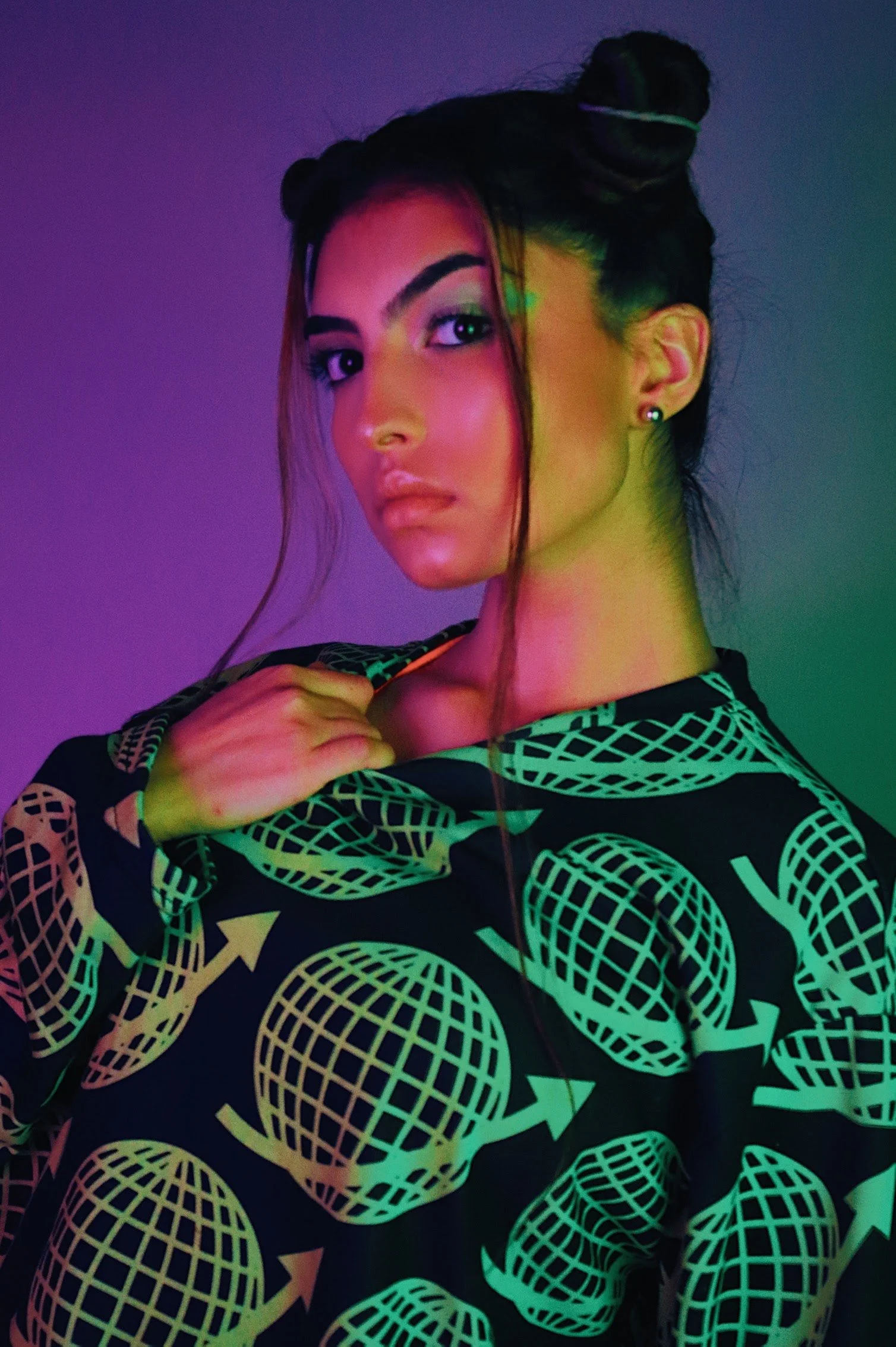 A woman with dark hair styled in two buns, wearing a black top with a green geometric pattern of globes and arrows, with neon-colored lighting affecting the appearance of the photo.