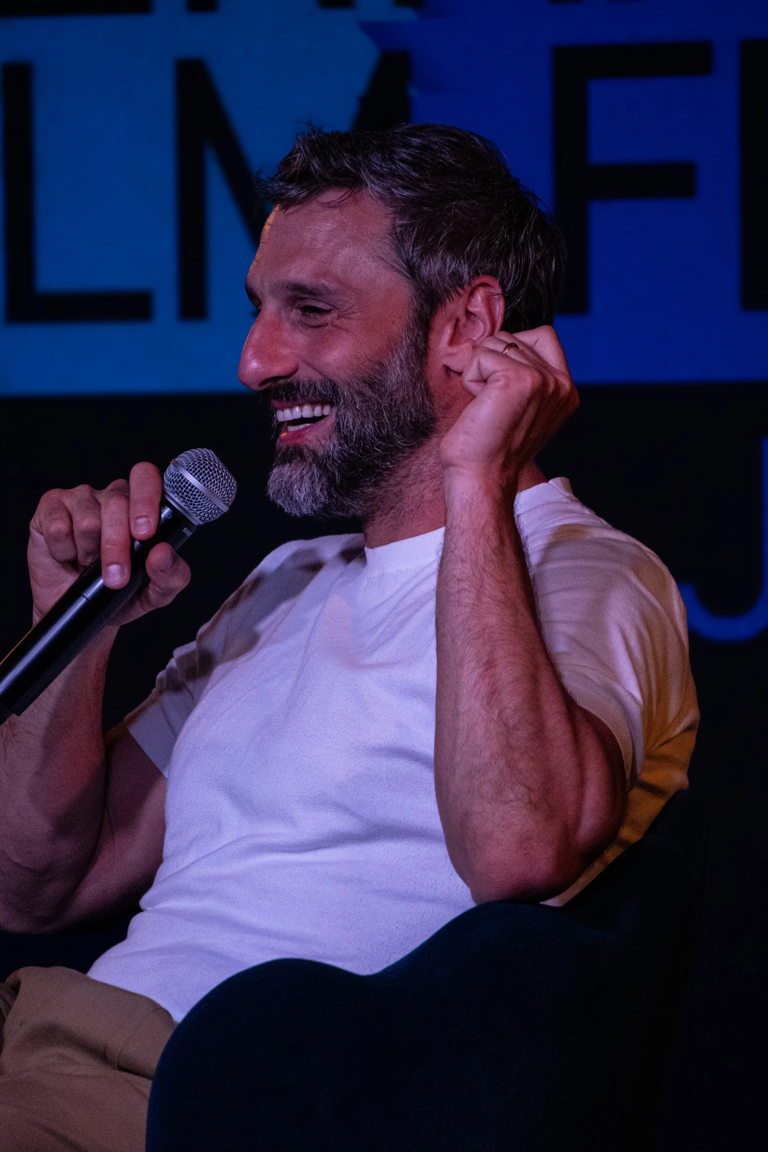 A man with a beard and short brown hair, wearing a white t-shirt, smiling and holding a microphone, appears to be speaking or singing on a stage with a blue background.