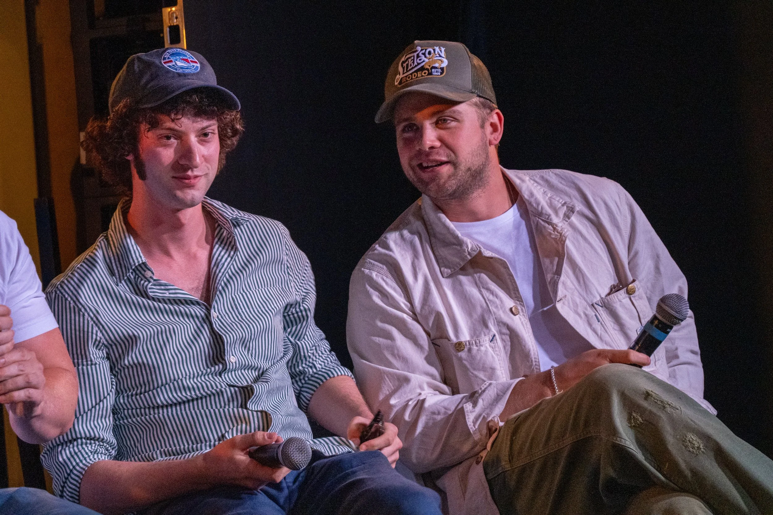 Two men sit on stage with microphones, one wearing a striped shirt and a black cap, the other in a white shirt and a gray cap, in a dark setting.