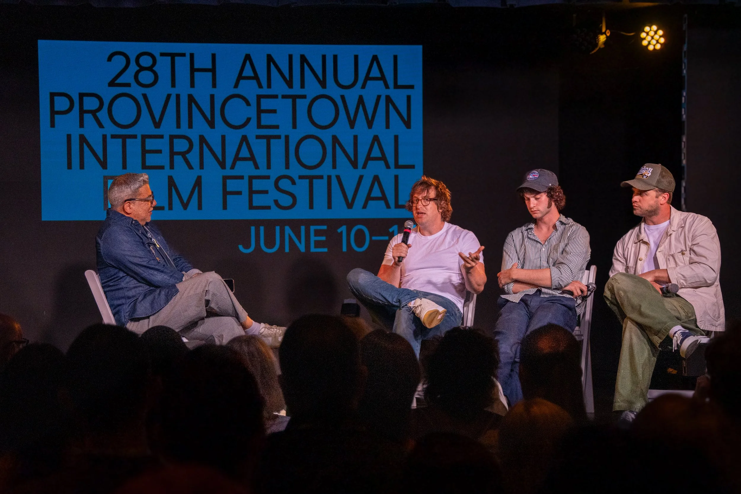 A panel discussion at the 28th Annual Provincetown International Film Festival held June 10-16. Four men are seated on stage, with one holding a microphone and speaking. A large blue sign with event details is in the background.