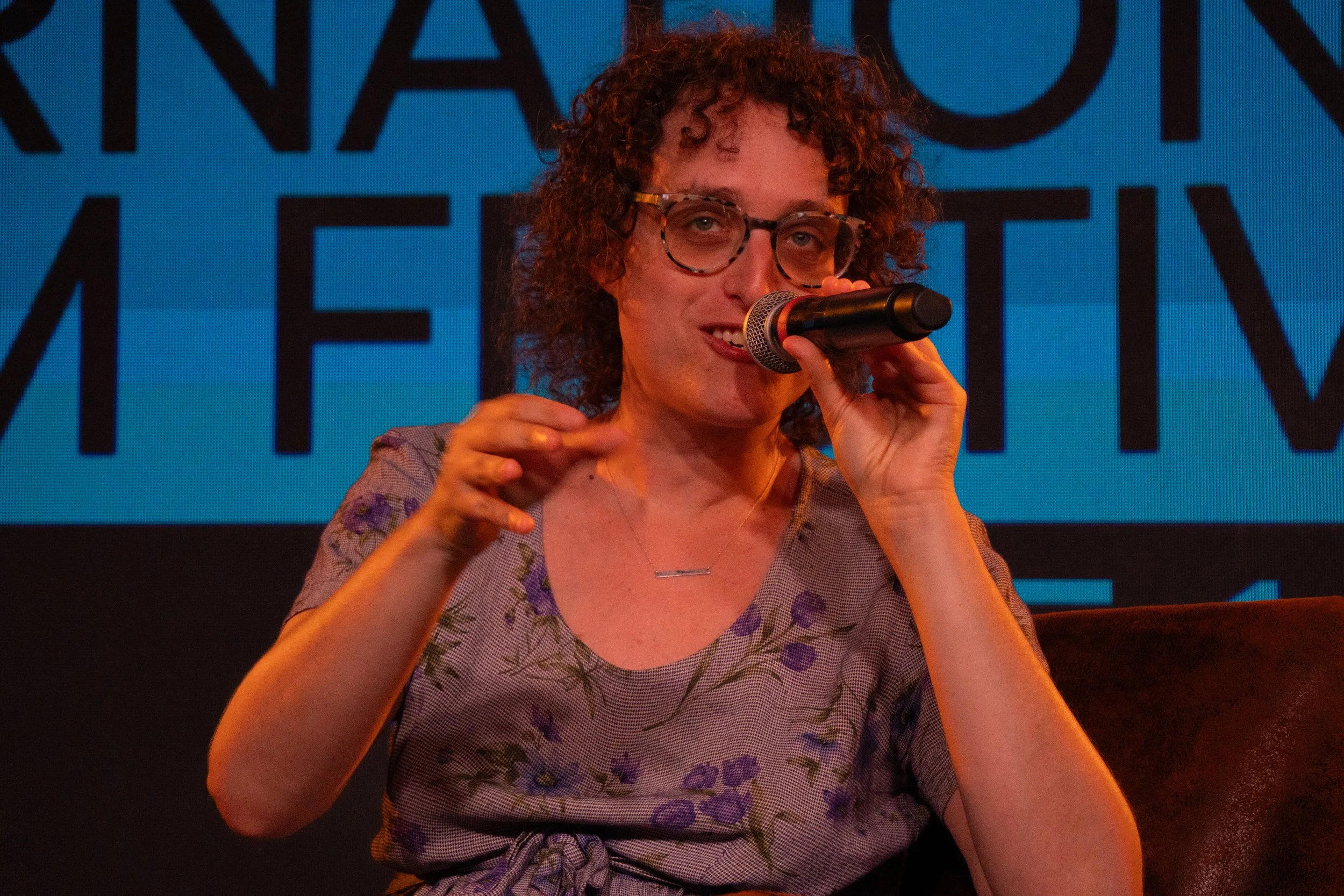 A woman with curly hair, glasses, and wearing a floral dress is speaking into a microphone at an event, with large text on a blue background behind her.