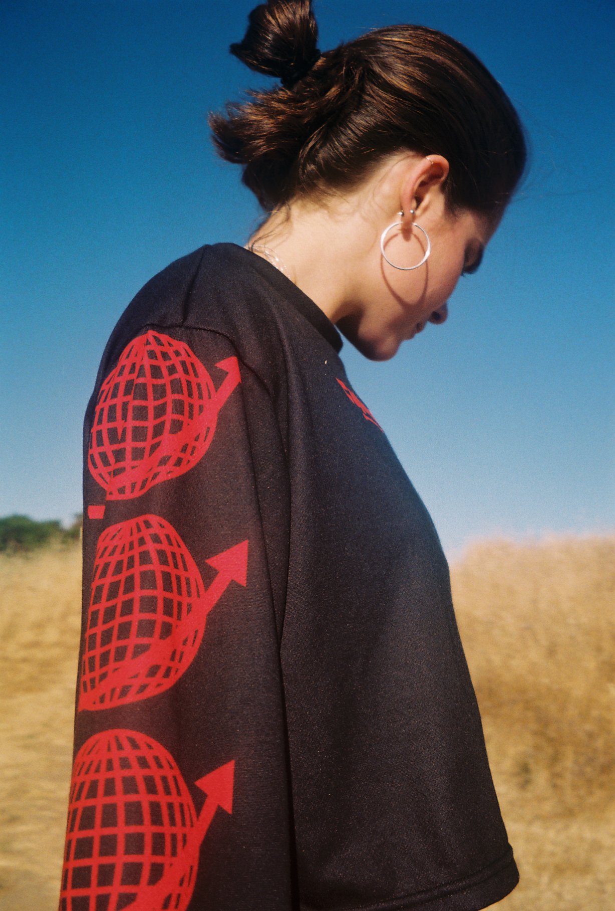 Side profile of a woman with brown hair tied up, wearing a black shirt with red globe and arrow designs on the sleeve, silver hoop earrings, against a clear blue sky and open field background.