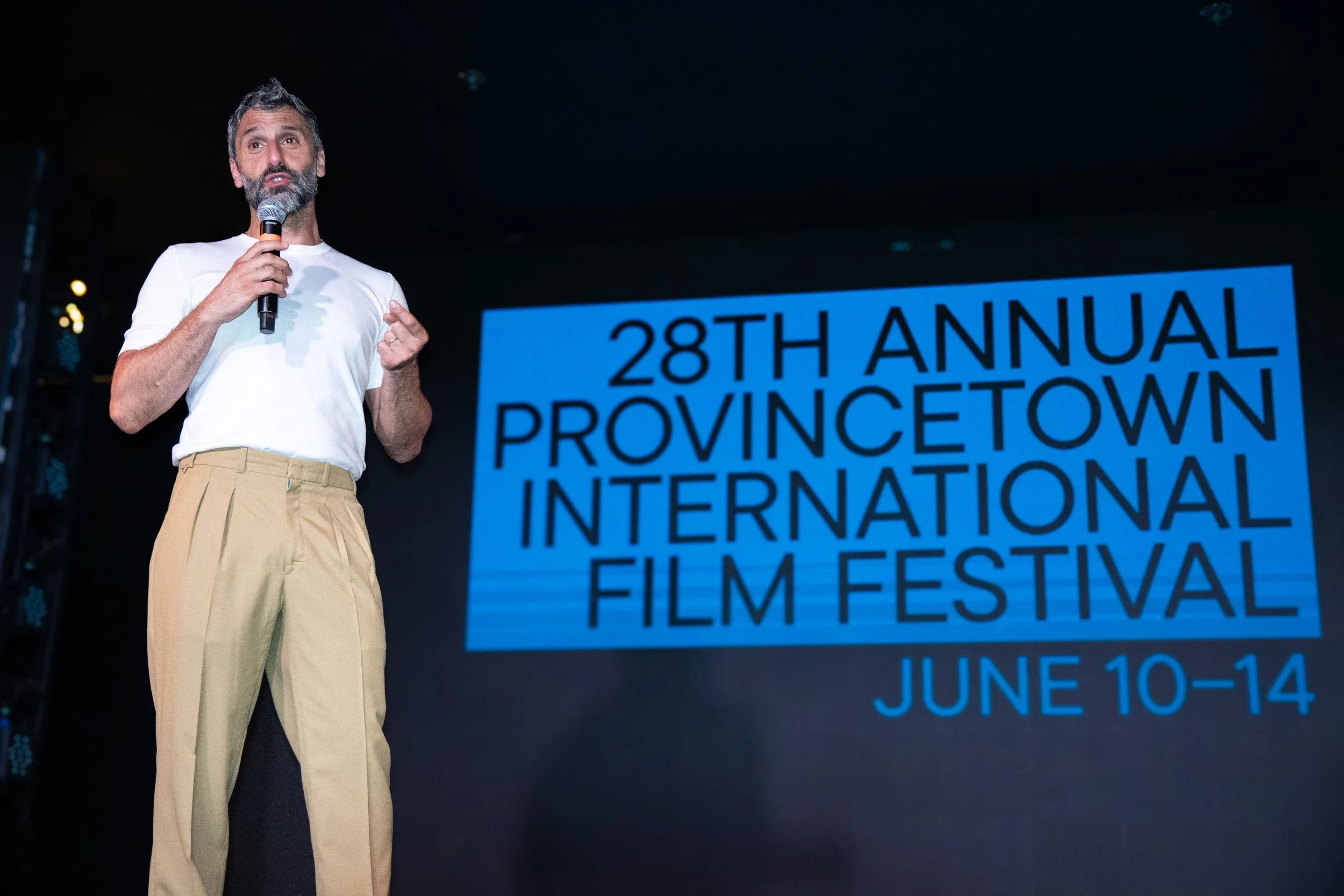 A man with gray hair and a beard holding a microphone, speaking on stage at the 28th Annual Provincetown International Film Festival, June 10-14.