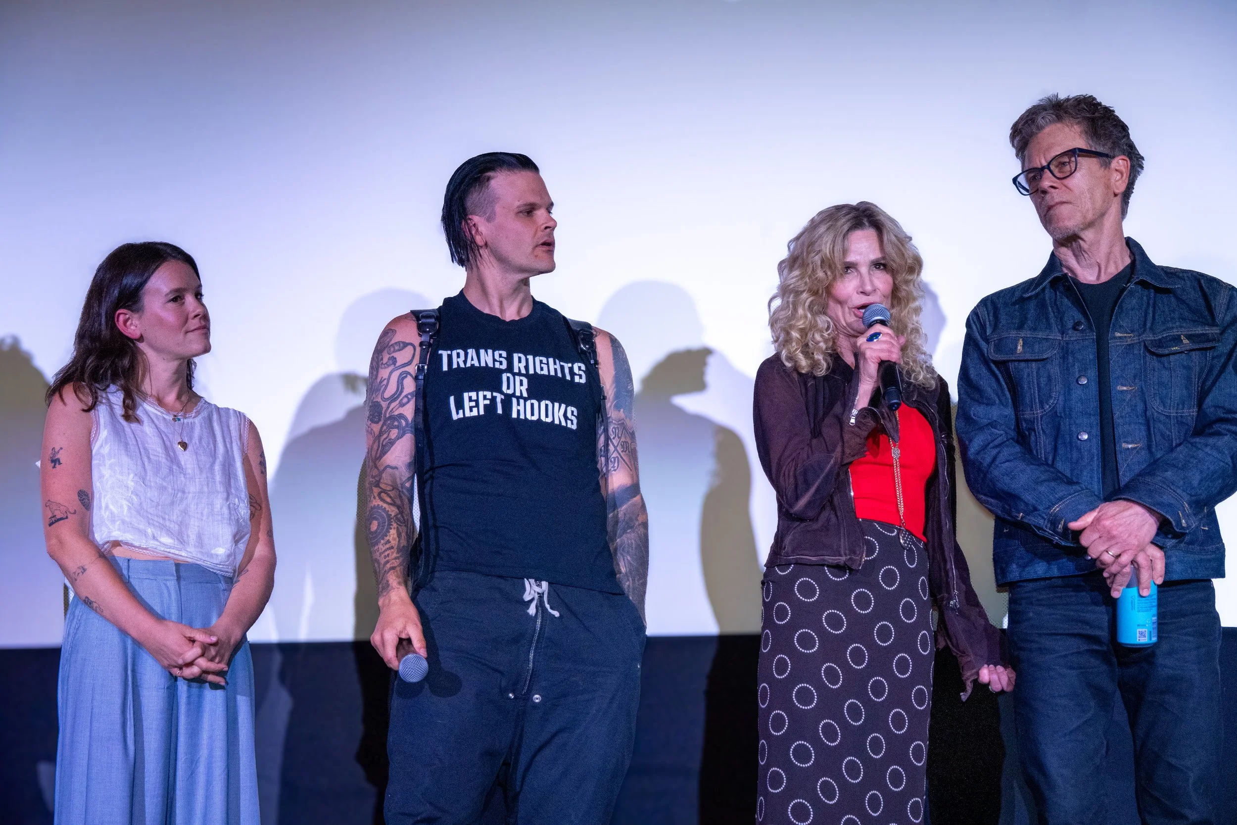 Group of five people standing on stage in front of a white screen, with one woman speaking into a microphone.