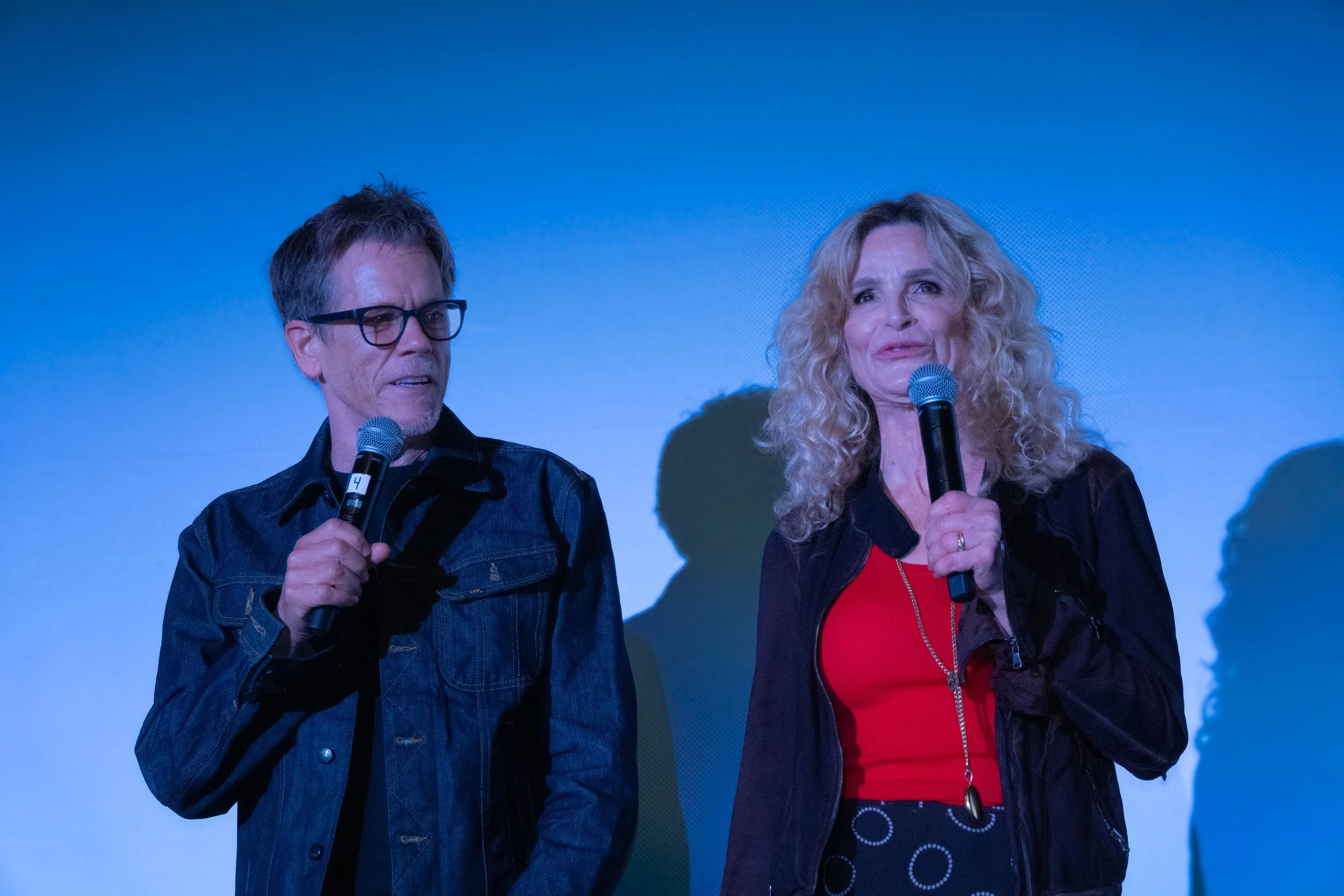 A man and woman are speaking into microphones on stage, with a blue background behind them.