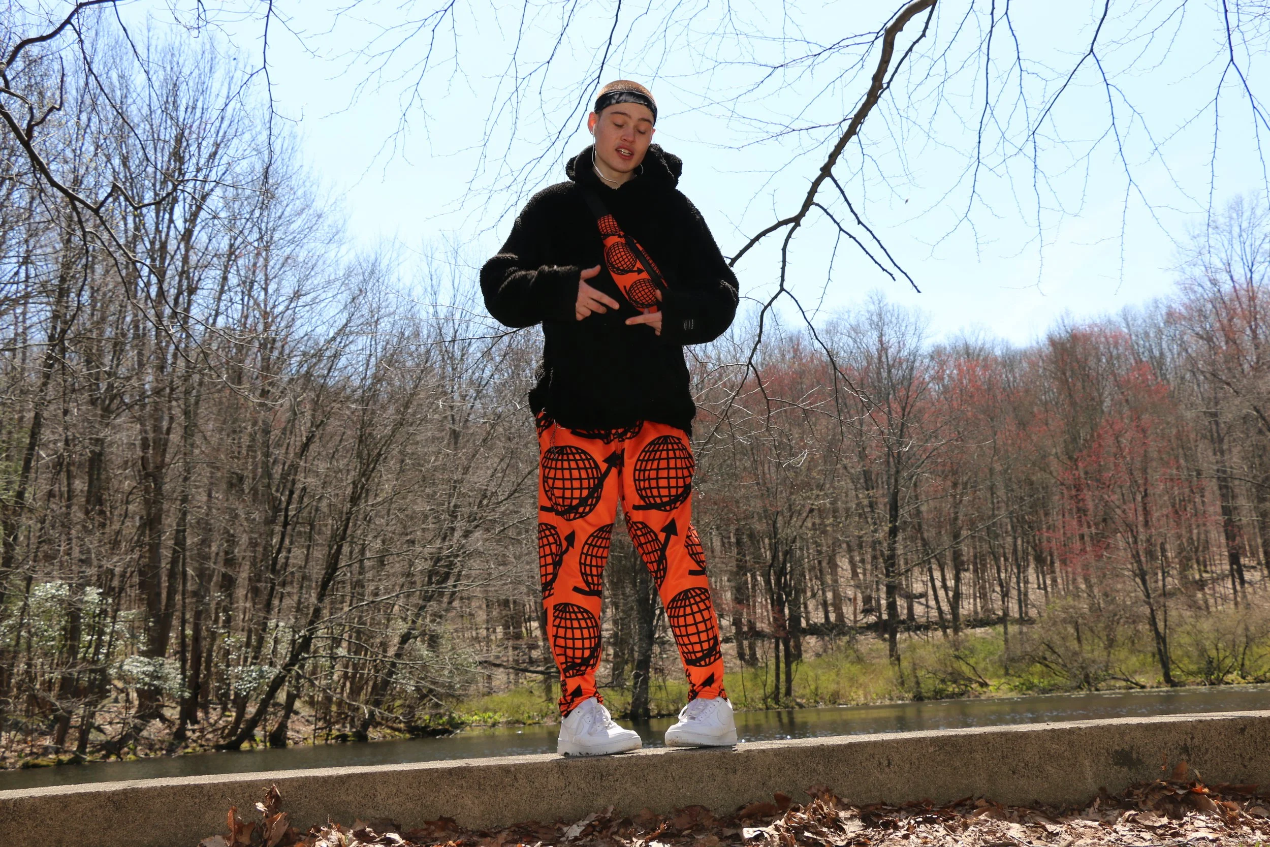 Young man wearing orange patterned pants, a black hoodie, and white sneakers standing on a concrete ledge in a wooded park during daytime with bare trees and a light blue sky.