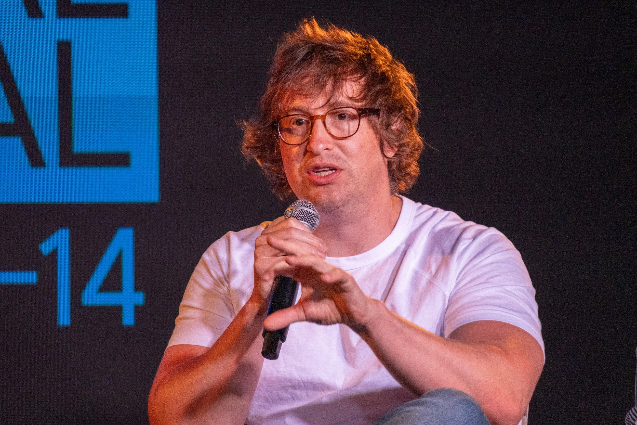 A man with curly hair and glasses wearing a white T-shirt, speaking into a microphone during an event.