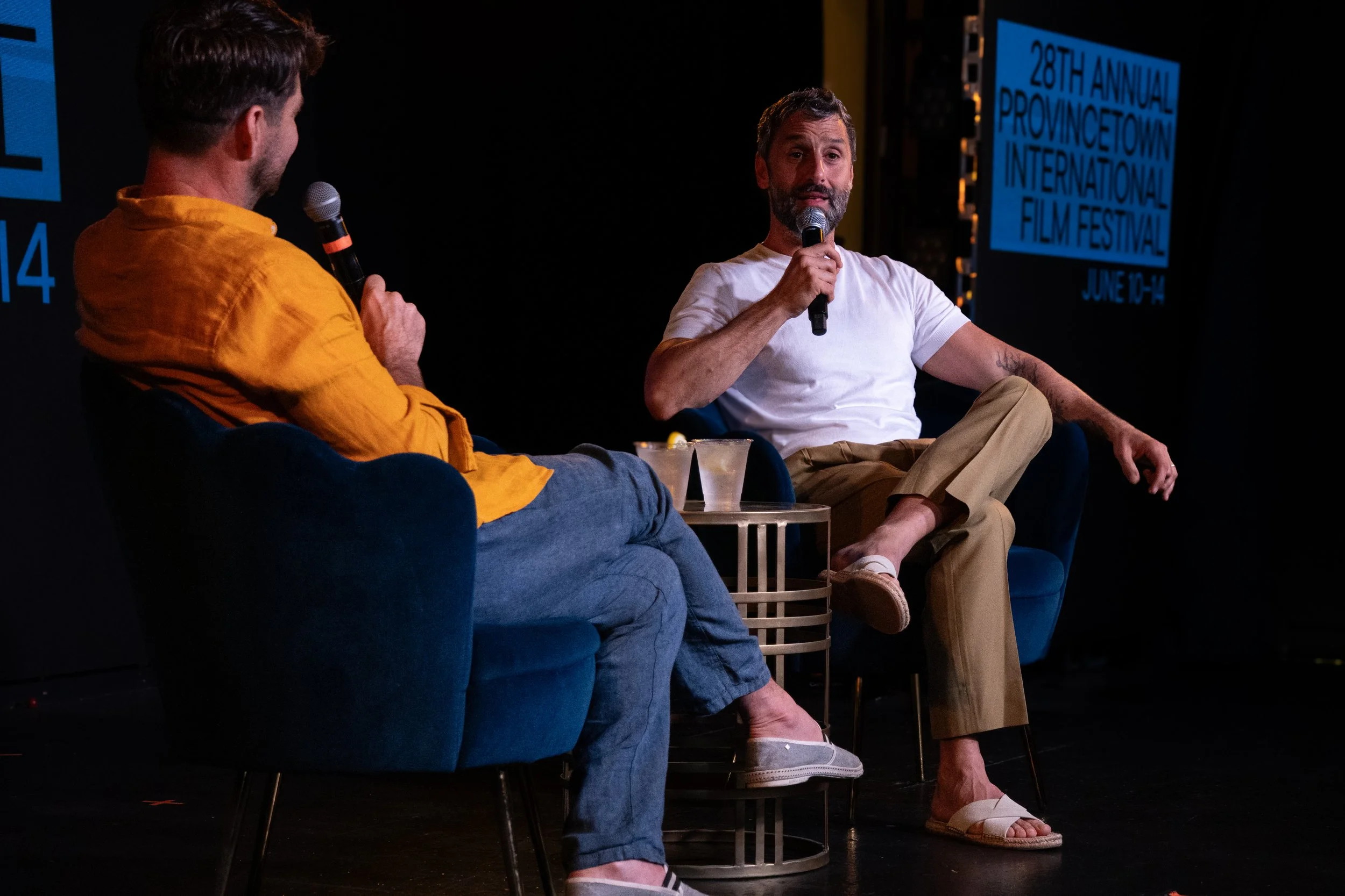 Two men are sitting on stage, each holding a microphone, engaged in a conversation during a panel event at the 28th Annual Provincetown International Film Festival. The man on the right is wearing a white t-shirt and beige pants, and the man on the l