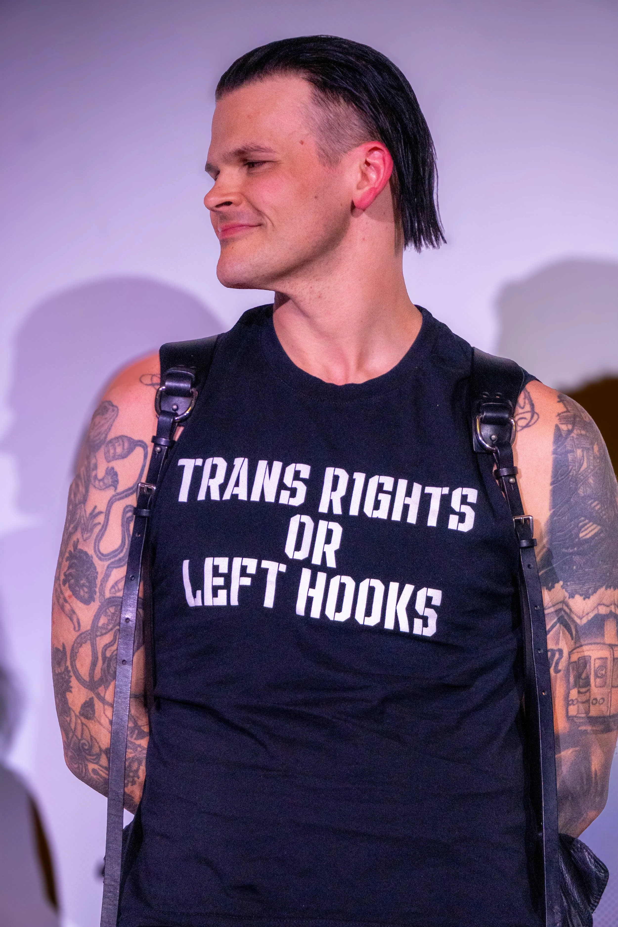 A person with a slicked-back hairstyle, wearing a black sleeveless shirt with a statement about transgender rights, and displaying tattoos on their arms. They are standing with a neutral background.