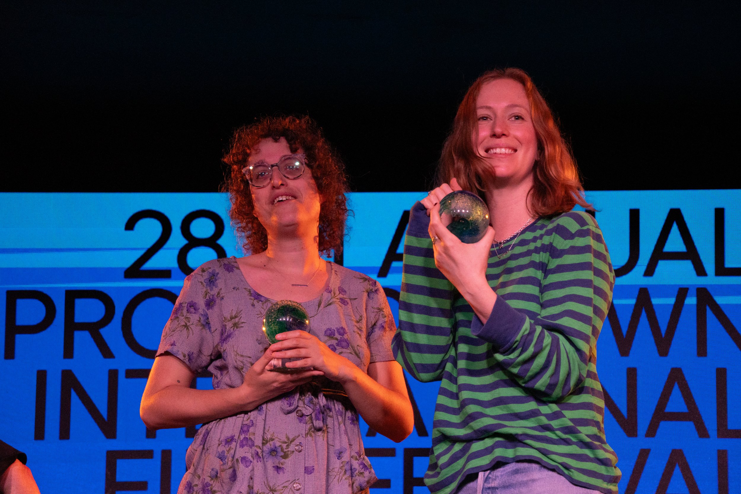 Two women standing on stage, holding trophies, smiling, with a blue background and some text, suggesting it is an award ceremony.