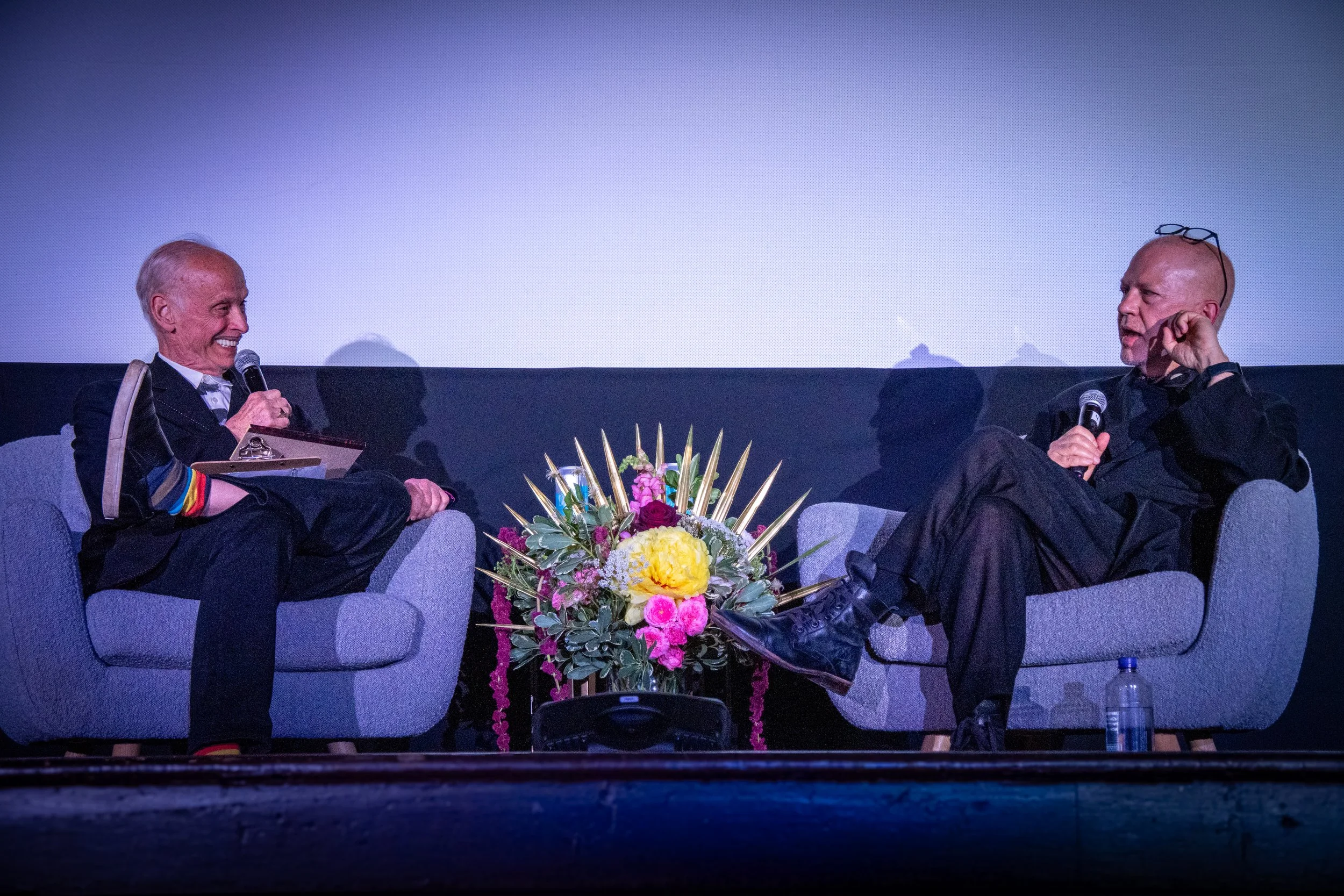 Two men sitting on stage with microphones, engaged in a conversation, with a floral arrangement between them and a blank screen behind them.