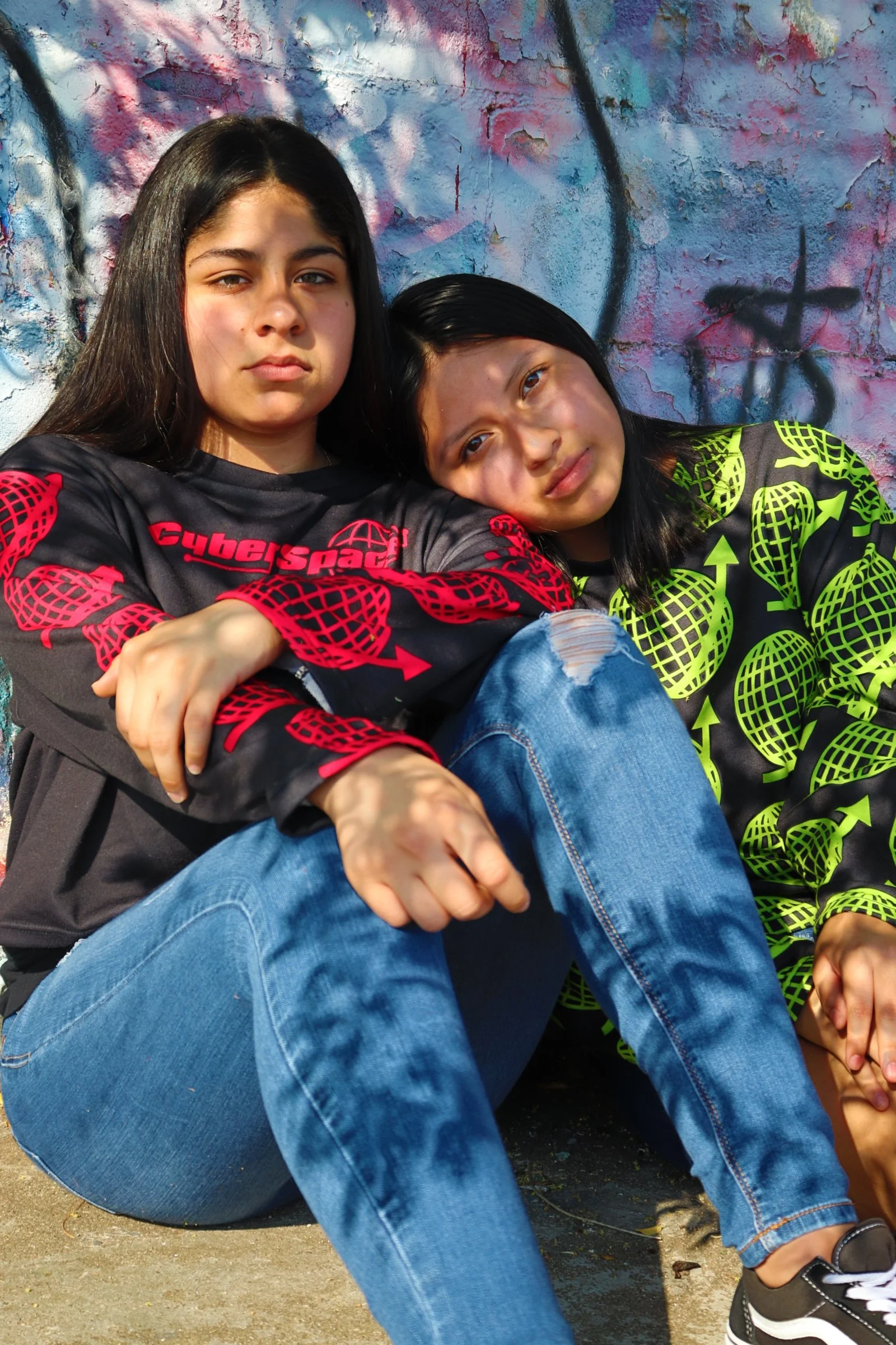 Two girls sitting on the ground against a colorful, graffiti-covered wall. The girl on the left has long dark hair and is wearing a black and red logo shirt, while the girl on the right has shoulder-length dark hair and is wearing a black and neon yellow patterned shirt. They are close together, with the girl on the right resting her head on the other's shoulder.