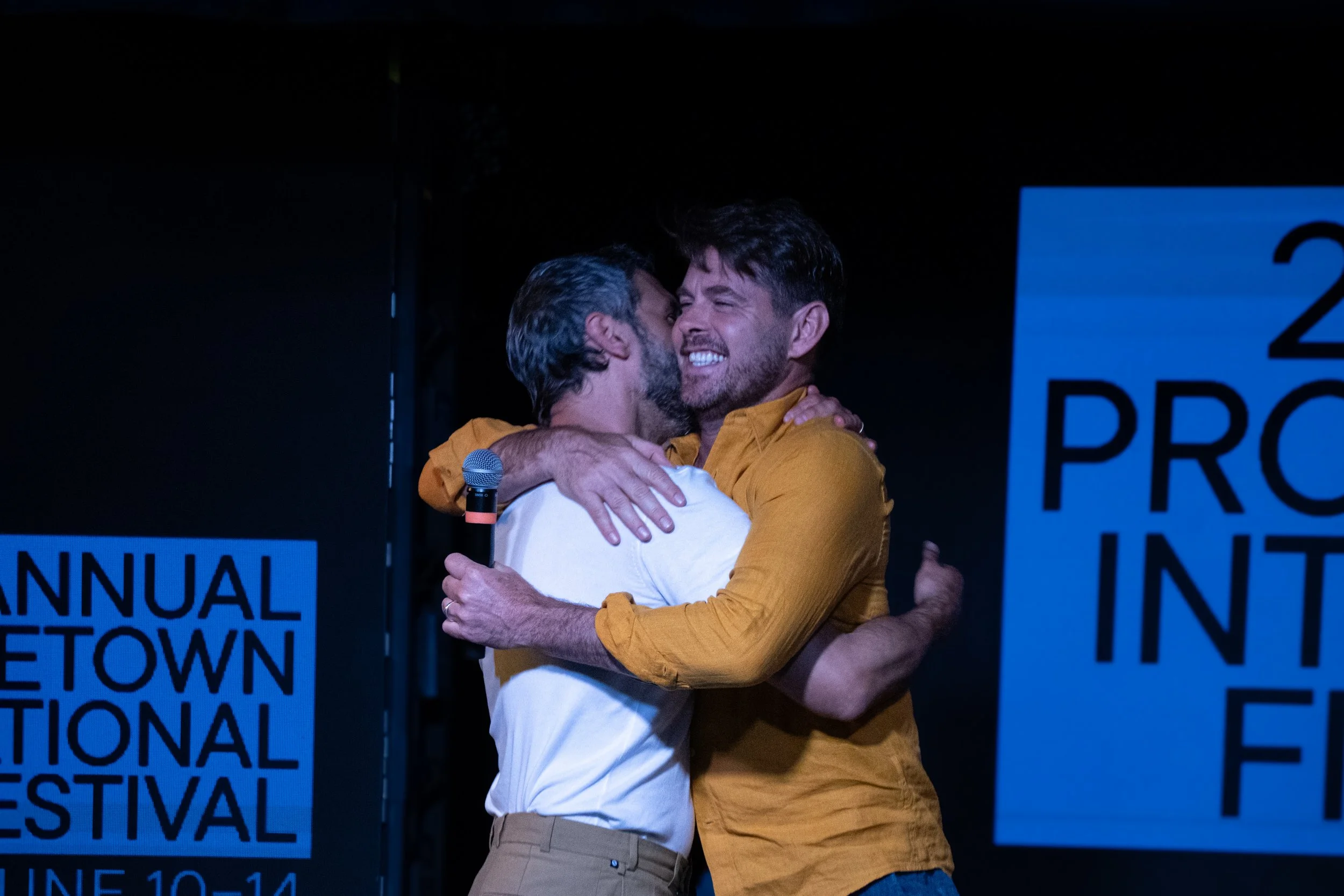 Two men hugging on stage at a festival, smiling and showing emotion, with the festival name partially visible in the background.