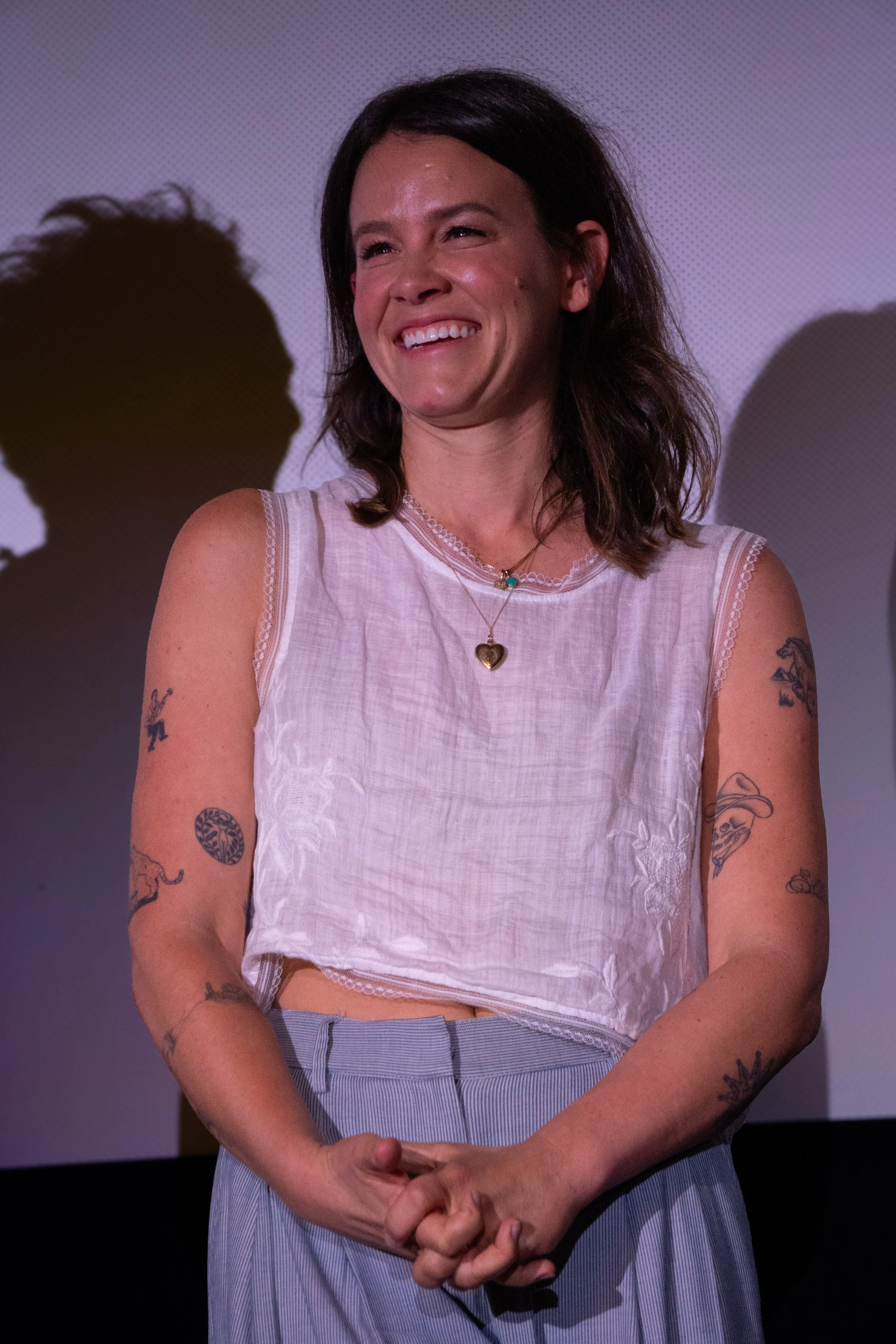 A woman with dark hair smiling, wearing a sleeveless white top and striped blue pants, with tattoos on her arms and necklaces around her neck, standing against a purple-lit background.