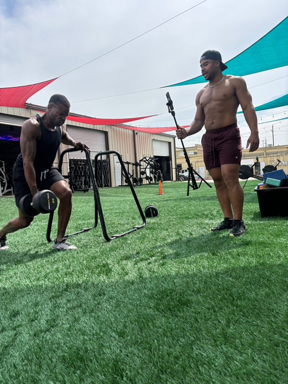 Two men exercising outdoors on artificial grass; one lifts a dumbbell while the other films with a camera on a pole.