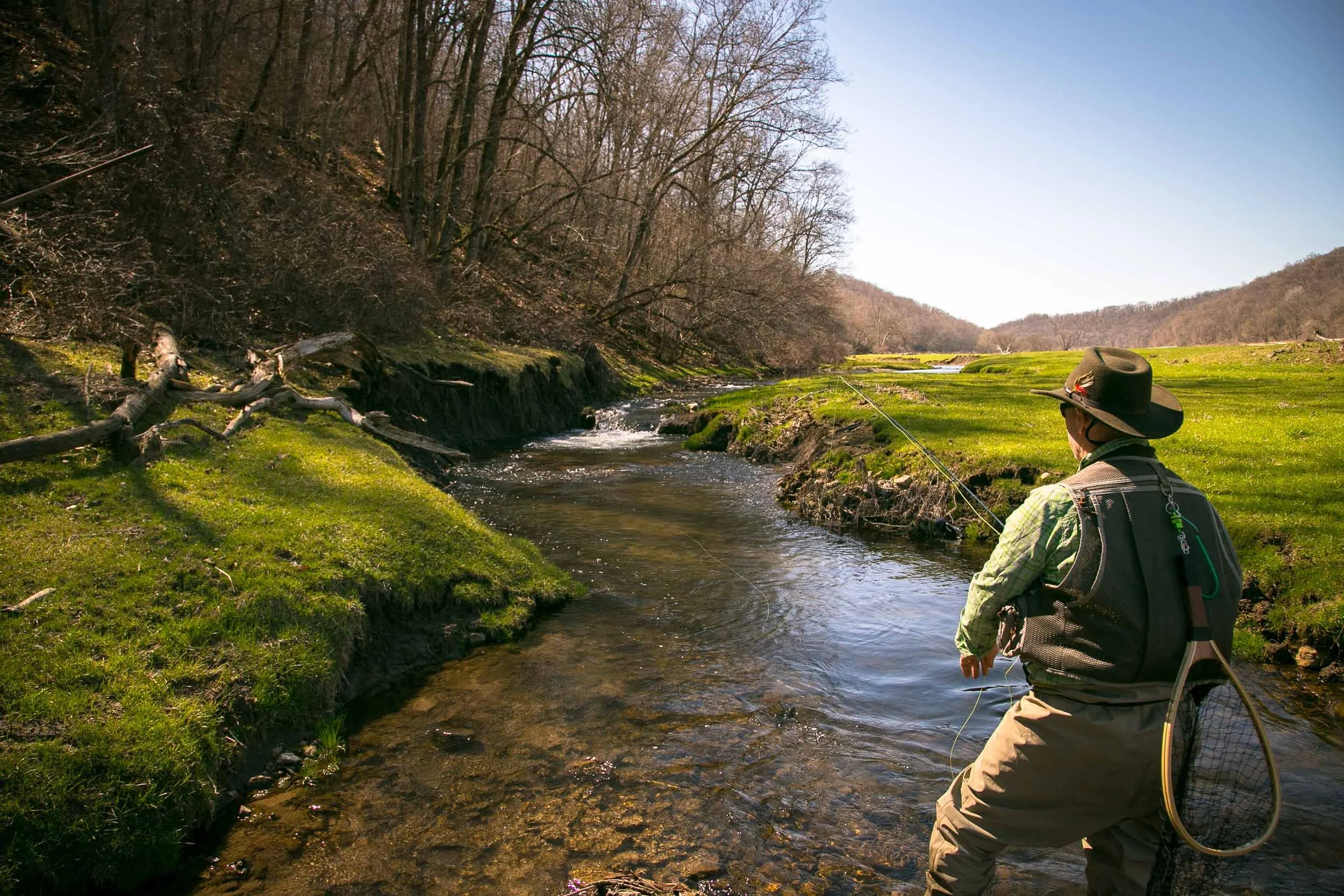 A person fishing in a river surrounded by green grass and trees on a sunny day.