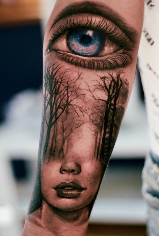 Surreal forearm tattoo combining a realistic blue eye, bare forest trees, and a woman’s face, by Colina.