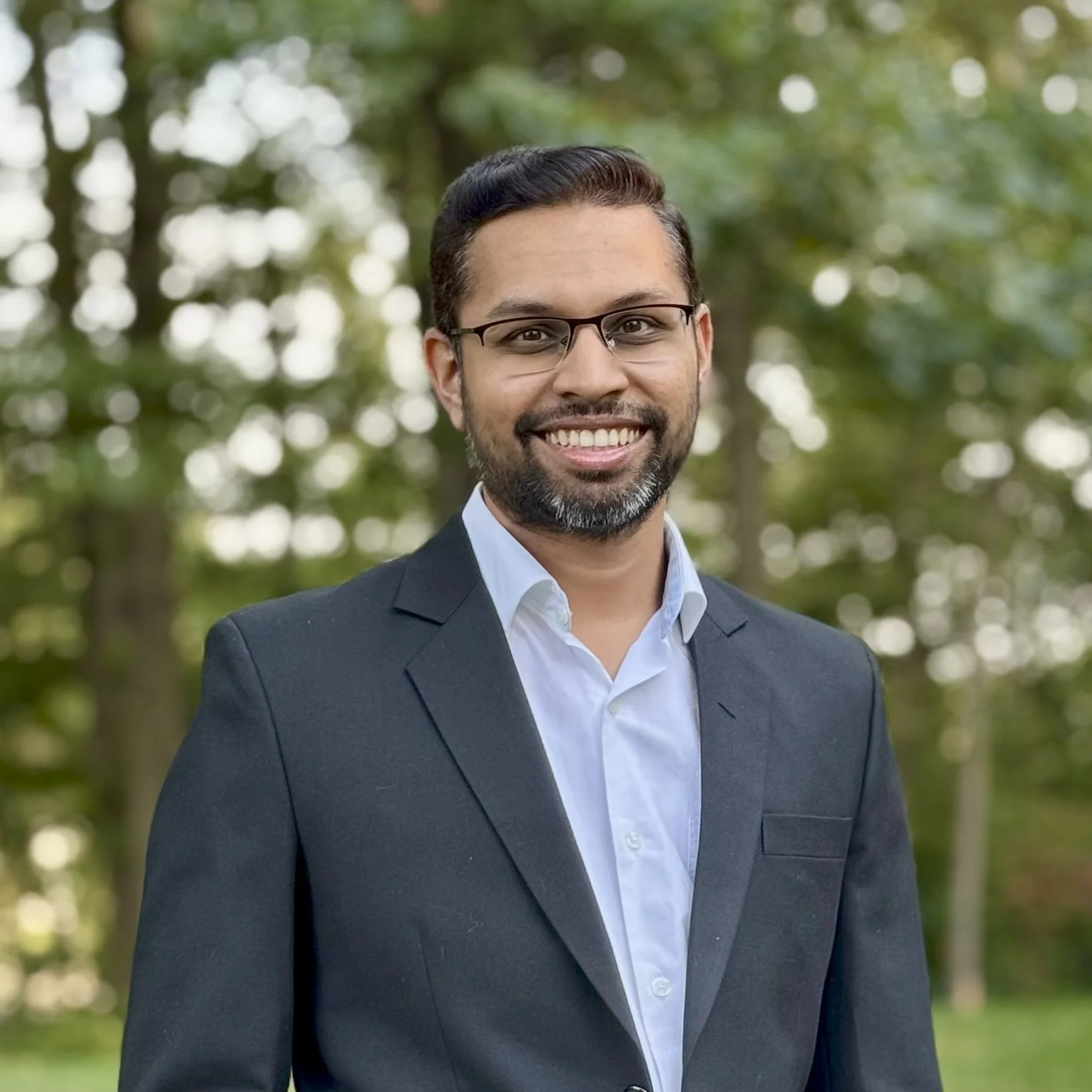 Portrait of a smiling man wearing a dark suit, glasses, and a white shirt, standing outdoors with a green, blurred background of trees.