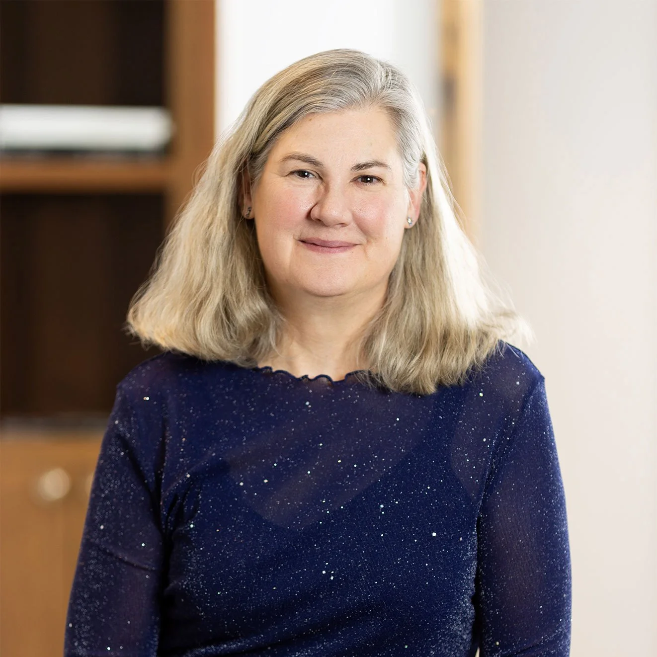 A middle-aged woman with shoulder-length blonde hair, wearing a dark blue, sparkly long-sleeved top, smiling gently, standing indoors near a bookshelf.