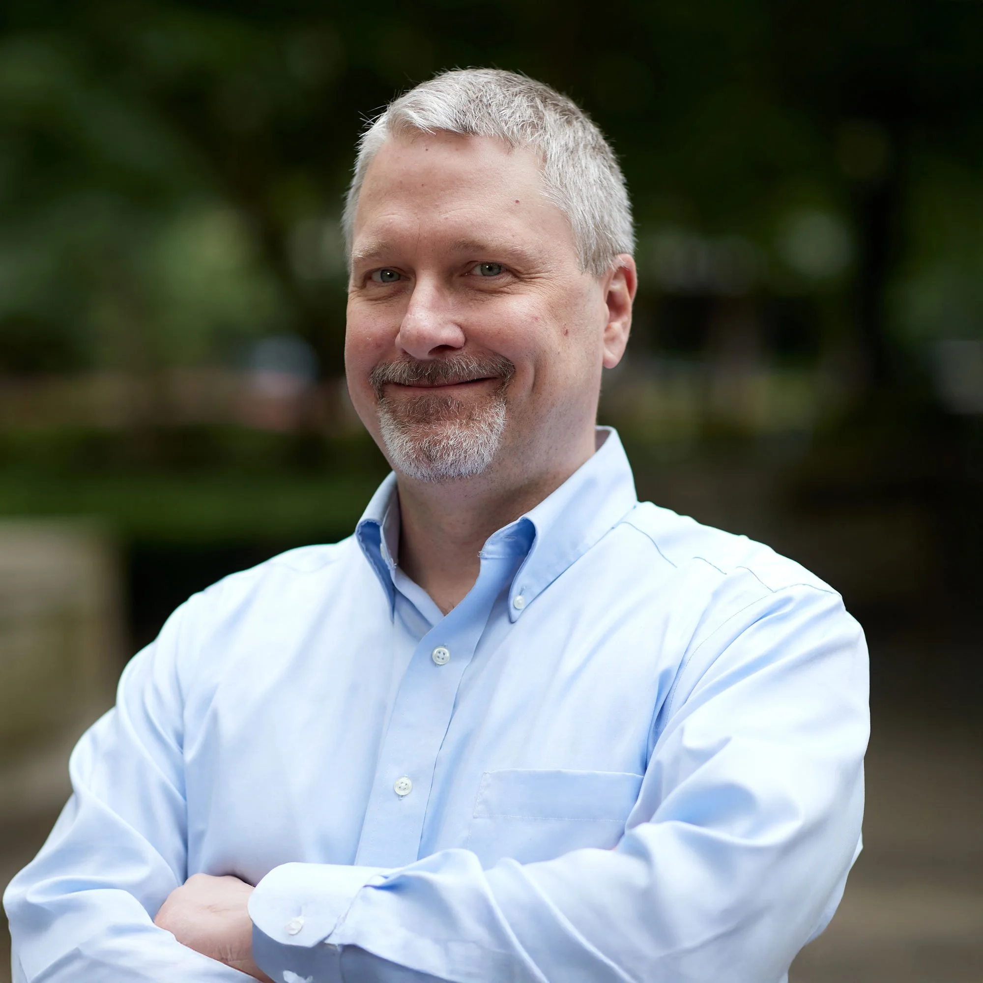 A middle-aged man with short gray hair and a beard, wearing a light blue dress shirt, standing outdoors with arms crossed, smiling at the camera.