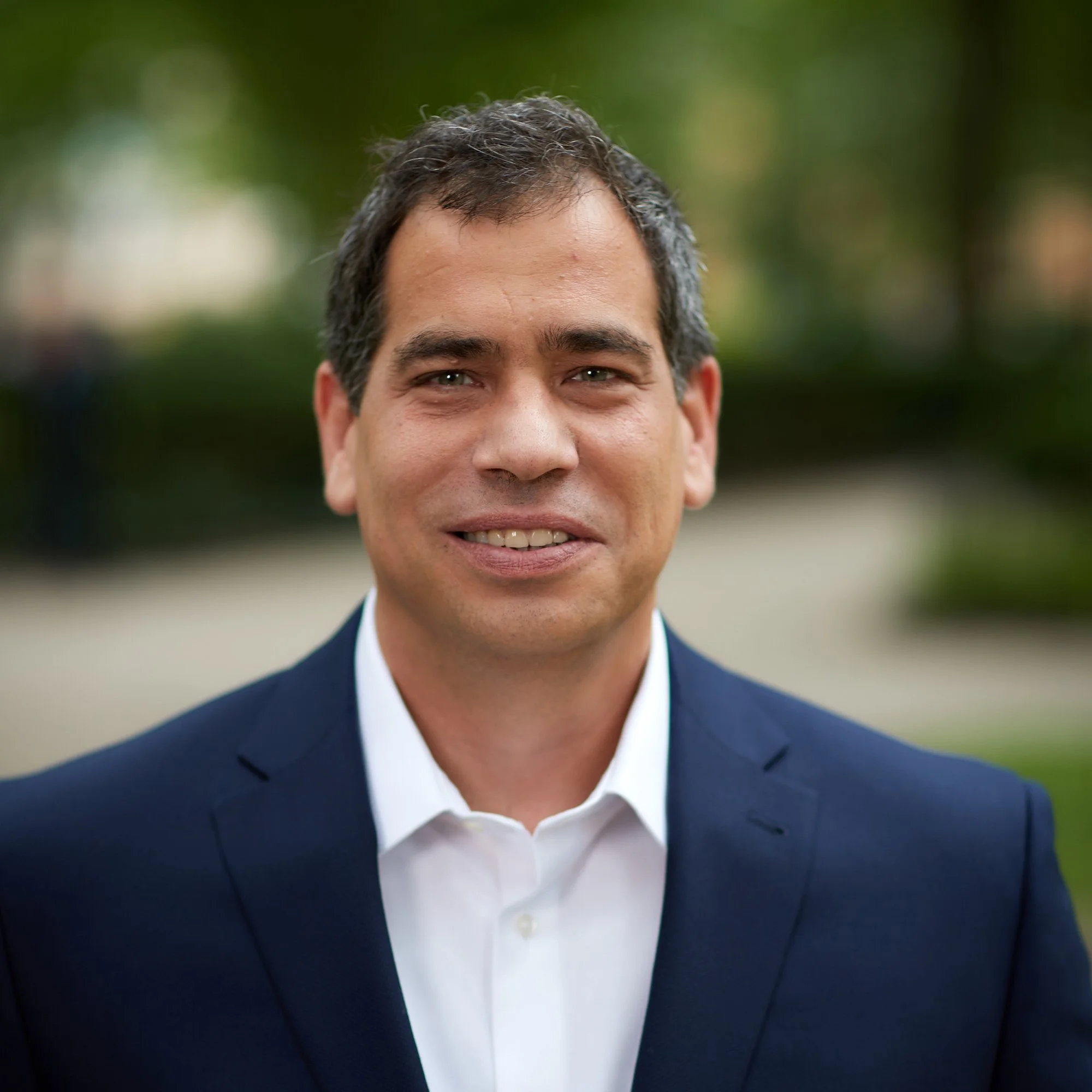 A man in a navy suit and white shirt smiling outdoors with blurred trees and pathways in the background.