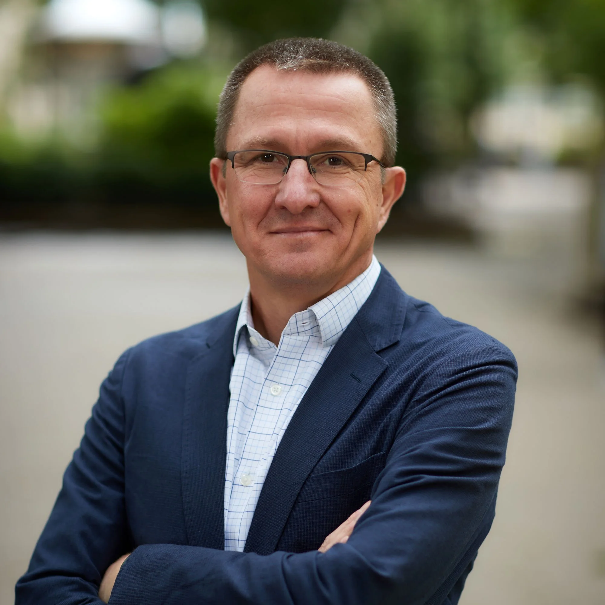 Portrait of a man with glasses, wearing a suit and crossing his arms, standing outdoors with blurred greenery in the background.
