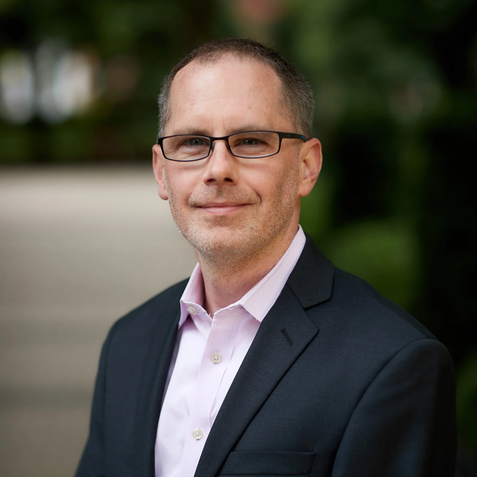 A man in a dark suit with a light pink shirt, wearing glasses, standing outdoors with blurred greenery in the background.