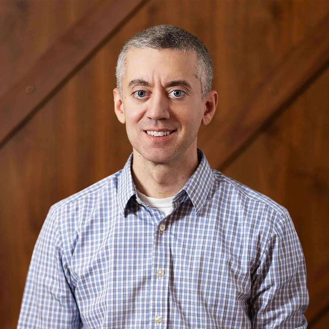 A man with short gray hair and blue eyes, wearing a plaid button-up shirt, standing in front of a wooden background.